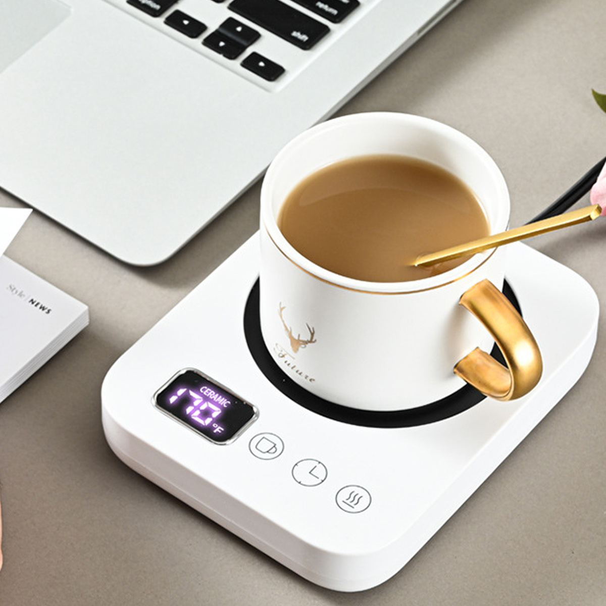Eine weiße Tasse mit Kaffee und goldenem Griff auf einem weißen Keramikwärmer. Ein Laptop und ein Notizblock sind ebenfalls sichtbar.