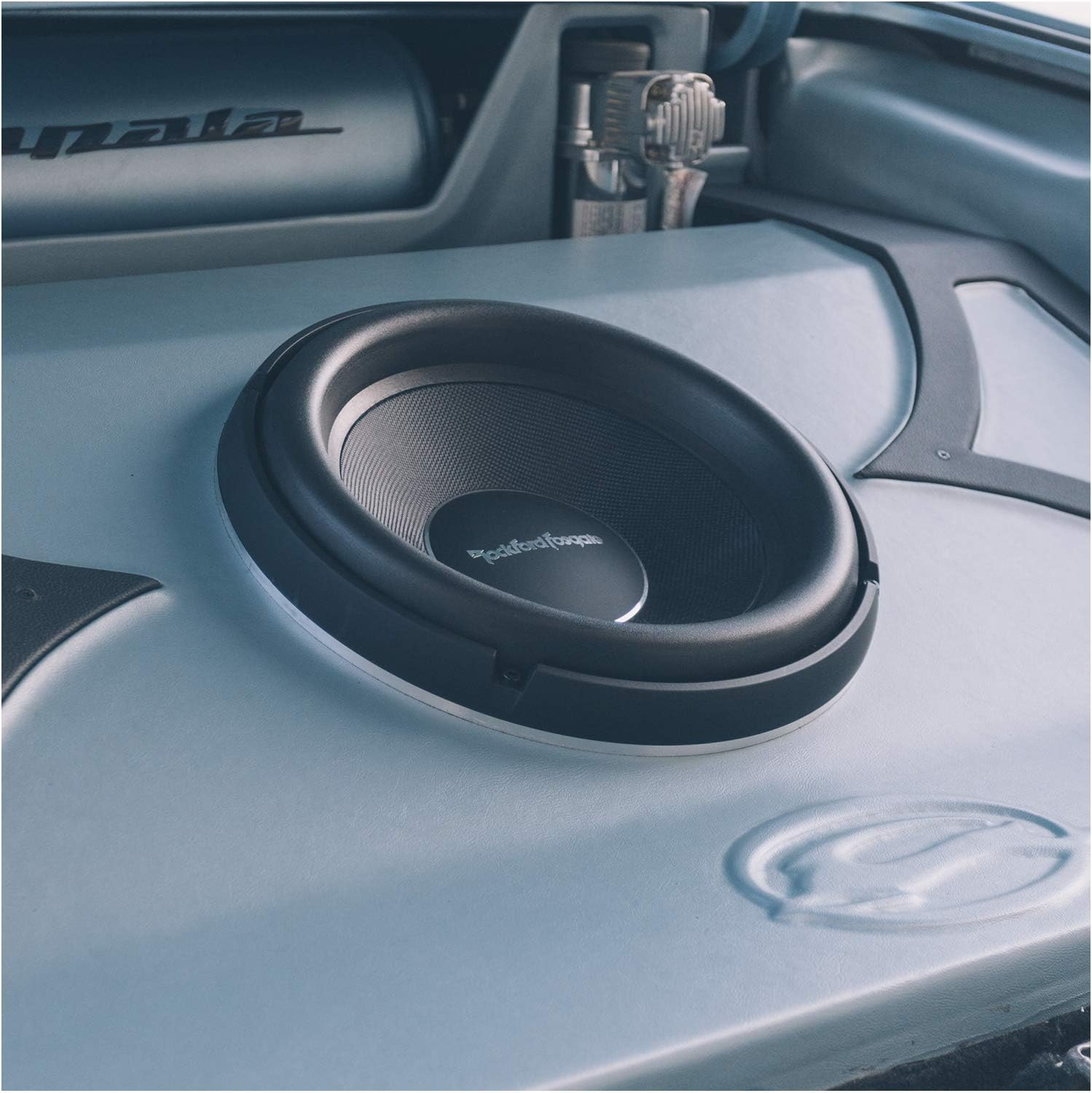 Der Kofferraum eines Autos zeigt einen großen schwarzen Subwoofer. Der Kofferraum ist hellgrau, im Hintergrund sind weitere Autoteile zu sehen.