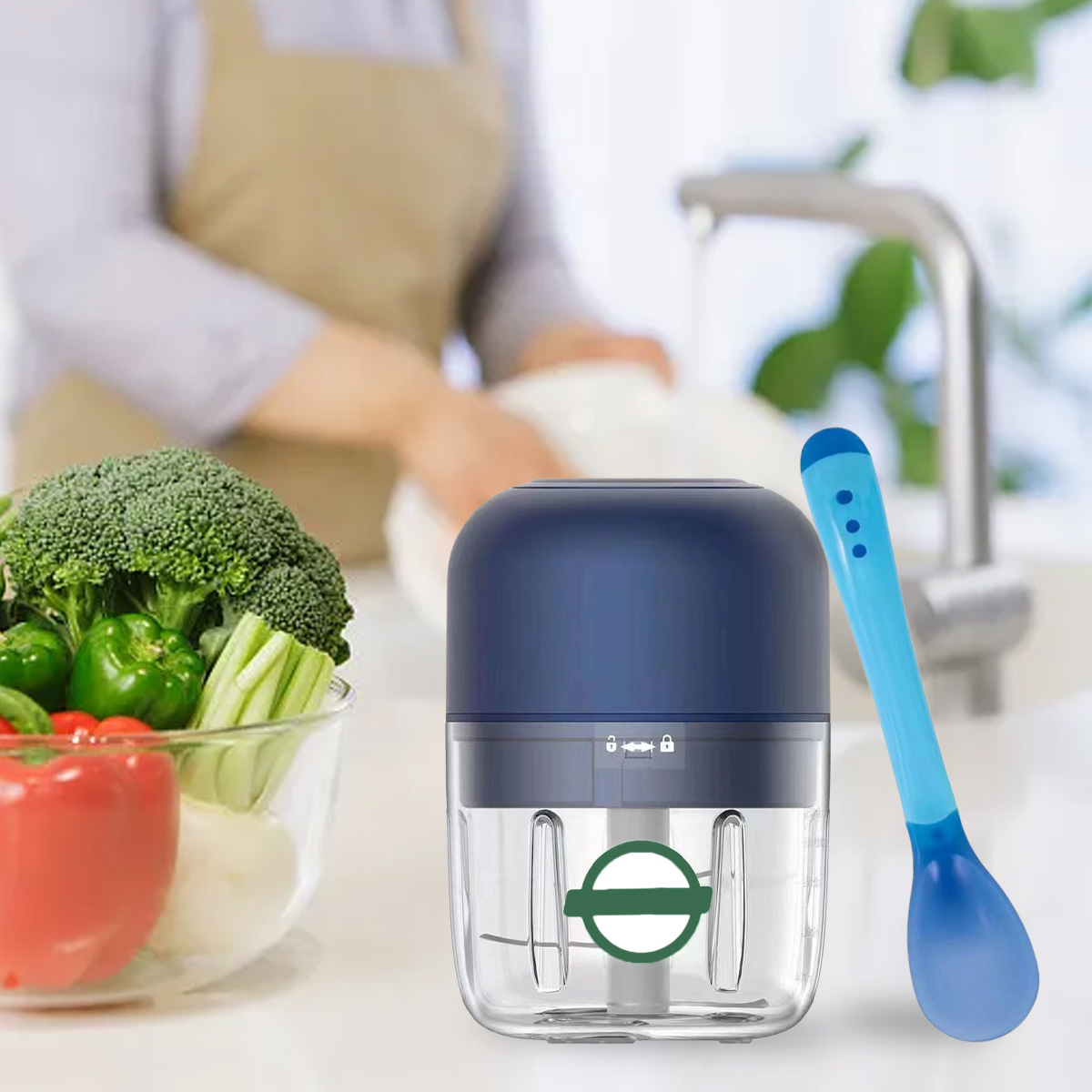 Küchenszene mit einem Foodprocessor, einem blauen Löffel und Gemüse in einer Schüssel. Im Hintergrund wäscht eine Person Geschirr.
