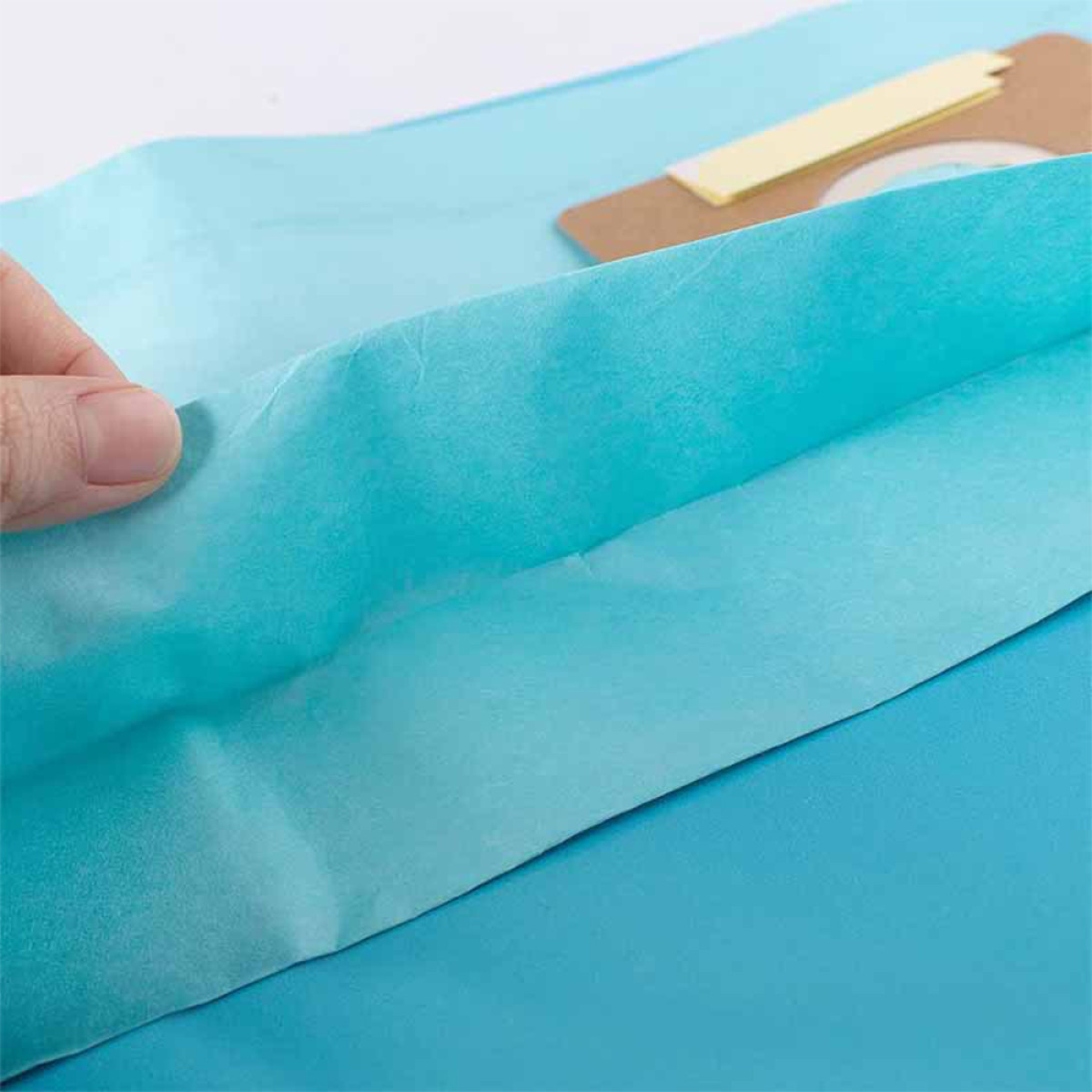 Eine Hand hält türkisfarbenes Seidenpapier hoch. Im Hintergrund hellblaues Papier und eine kleine Karte mit gelbem Tab.