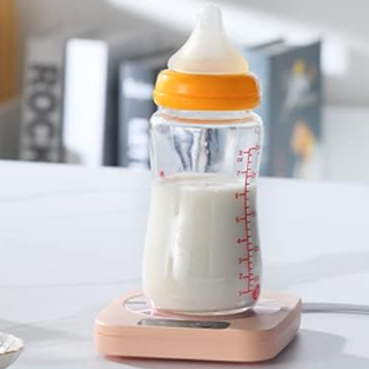 Eine Babyflasche mit Milch steht auf einem rosa Wärmer. Die Flasche ist durchsichtig mit gelbem Ring und Sauger. Im Hintergrund Bücher.