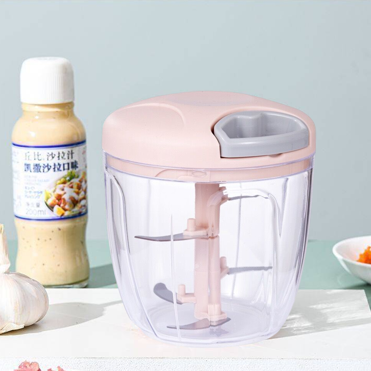 Ein rosa manueller Zerkleinerer mit Klingen, eine Salatdressingflasche und eine Knoblauchzehe auf einer weißen Oberfläche.