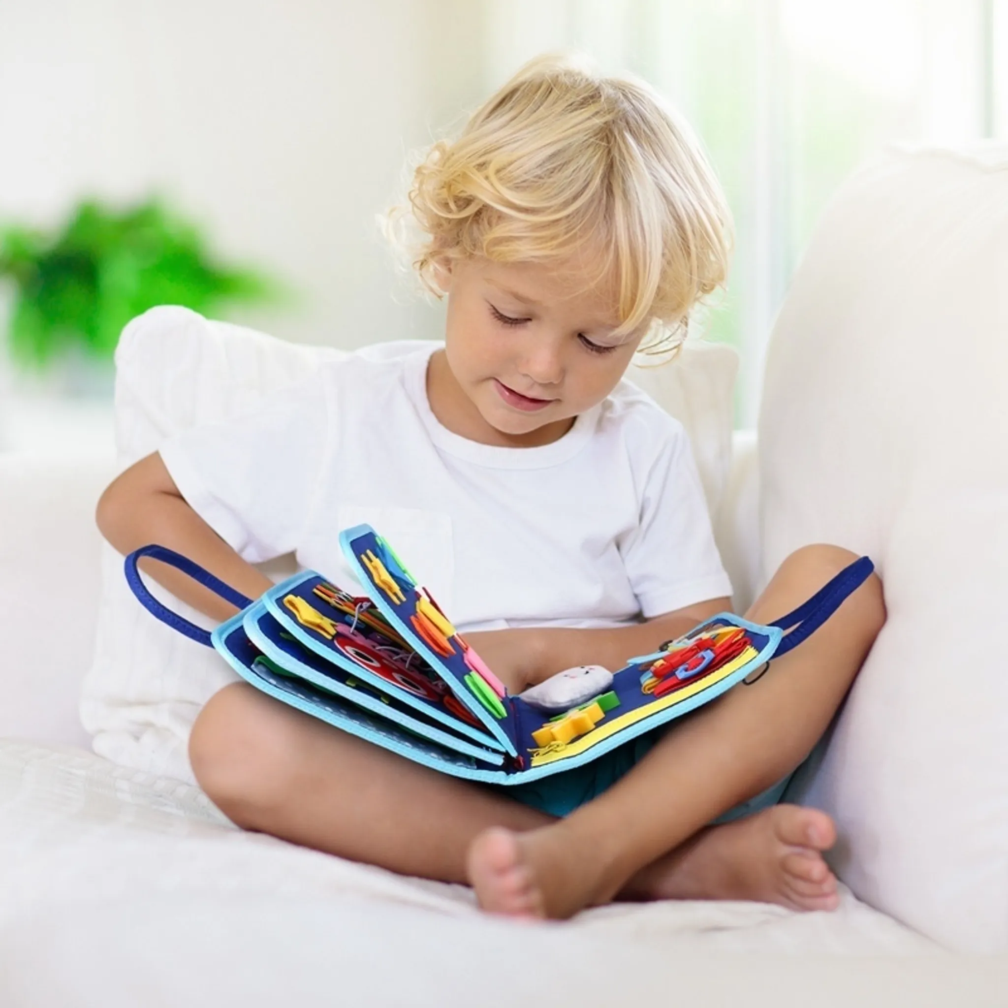 Ein Kind mit blonden Haaren sitzt auf einem weißen Sofa und betrachtet ein farbenfrohes Aktivitätsbuch. Der Hintergrund ist unscharf.
