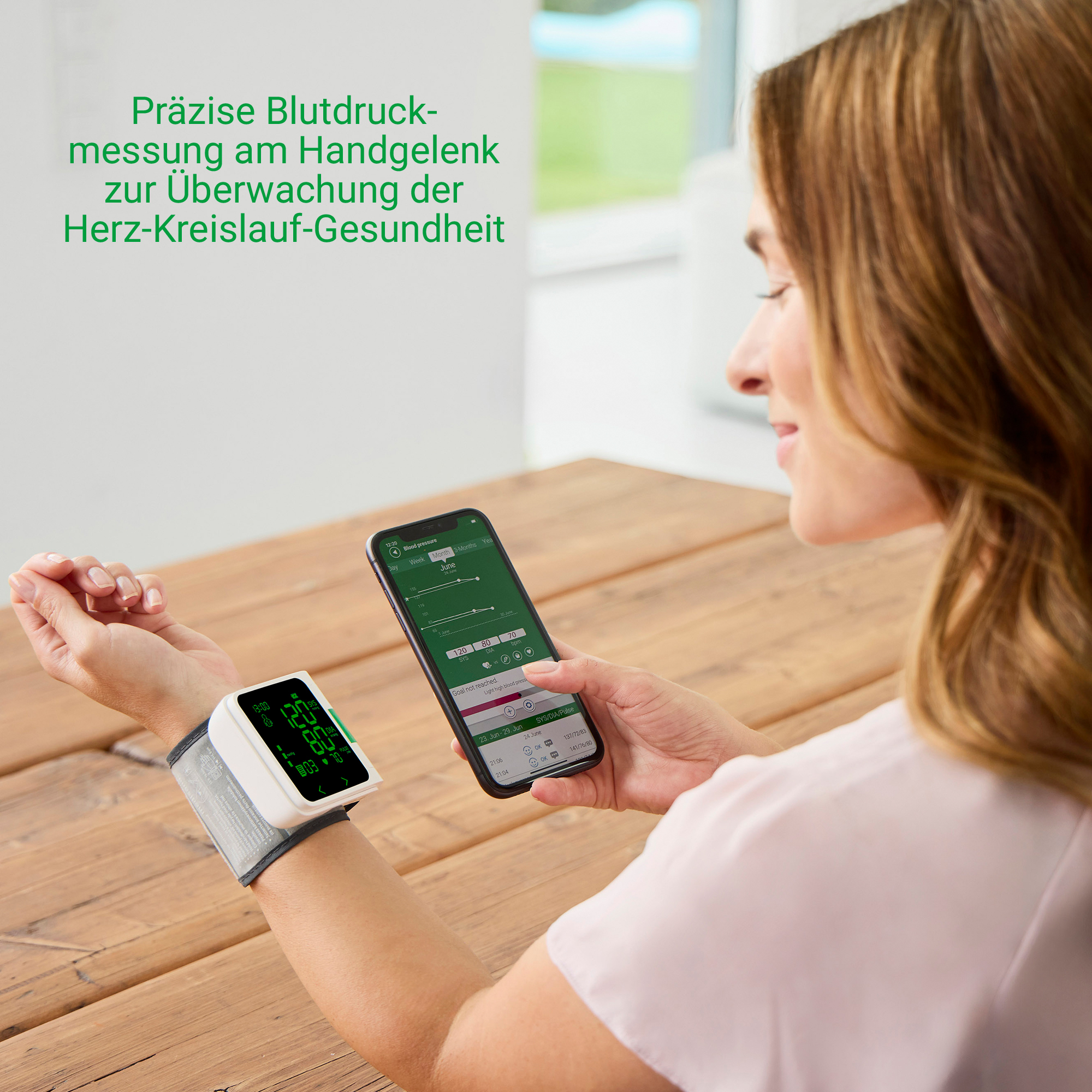 Eine Frau misst mit einem Gerät den Blutdruck am Handgelenk. Ein Smartphone zeigt die Messwerte auf einem Holztisch.