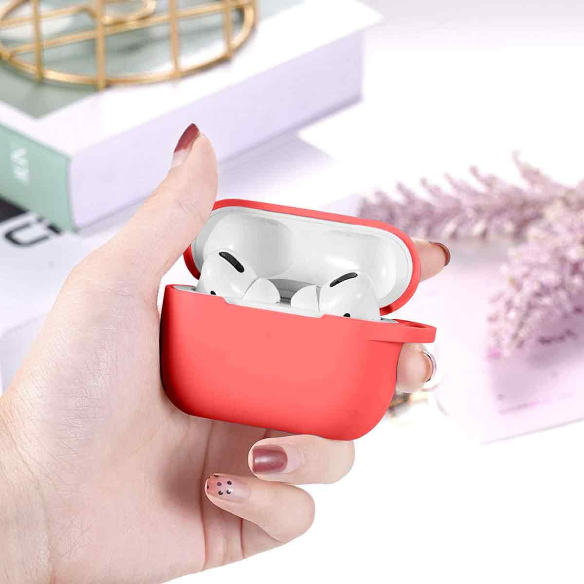 Eine Hand hält eine offene rote AirPods-Hülle, die weiße Ohrhörer zeigt. Im Hintergrund befinden sich Dekorationen und ein Buch.