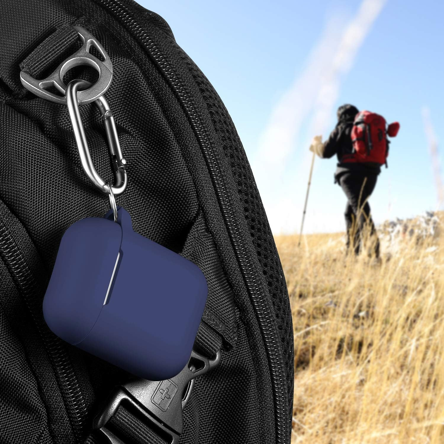 Ein blaues AirPod-Etui ist an einem schwarzen Rucksack befestigt. Eine Person wandert im Hintergrund mit rotem Rucksack an einem sonnigen Tag.