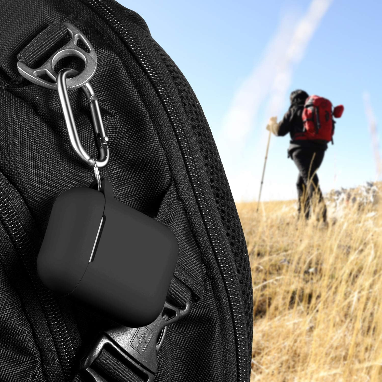 Schwarzes AirPods-Etui mit Karabiner an einem Rucksack befestigt. Im Hintergrund wandert ein Wanderer mit rotem Rucksack auf einem Hügel.