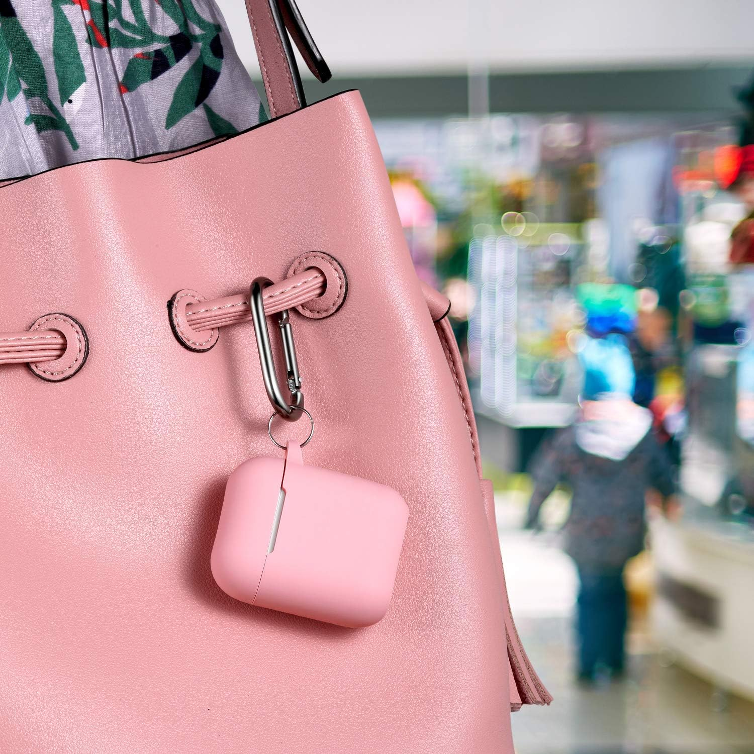 Eine rosa Handtasche mit angebrachtem rosa AirPods-Case hängt. Der Hintergrund ist eine verschwommene Ansicht eines Geschäfts mit einem Kind.
