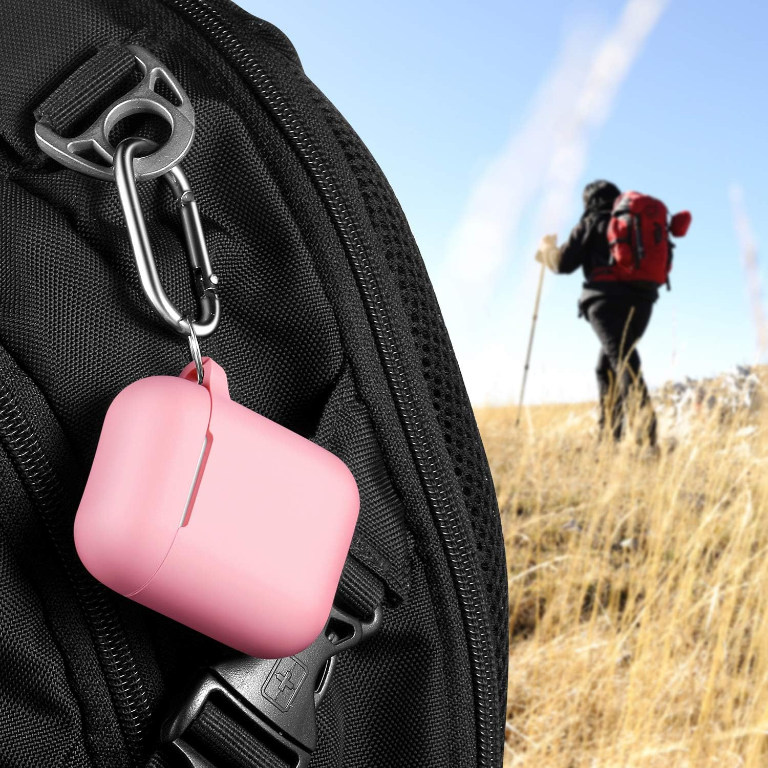 Rosa AirPods-Hülle an einem schwarzen Rucksack befestigt. Wanderer im Hintergrund mit rotem Rucksack beim Wandern in einem Feld.