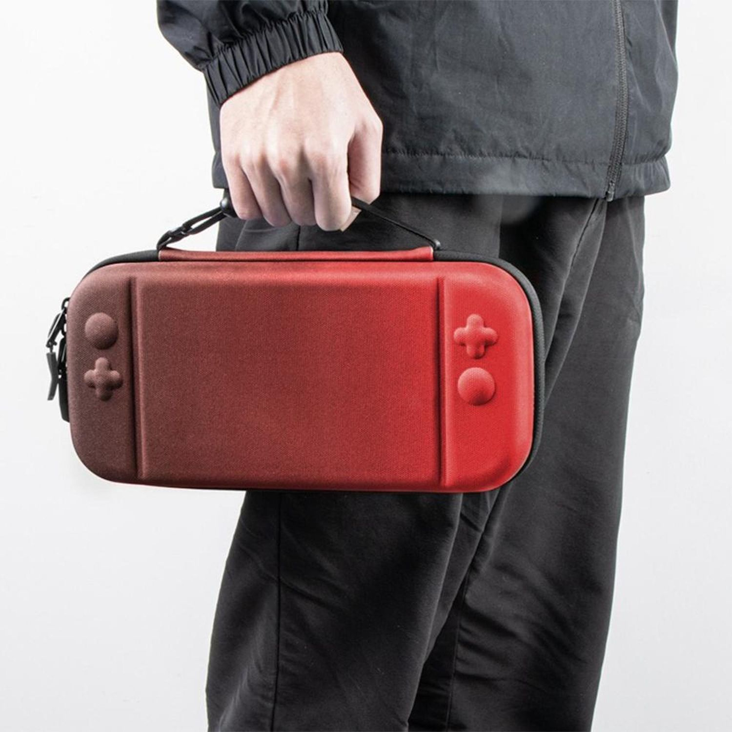 Una persona sostiene una funda roja y negra de Nintendo Switch. La funda tiene botones de control y la sujeta una mano. La persona viste ropa negra.