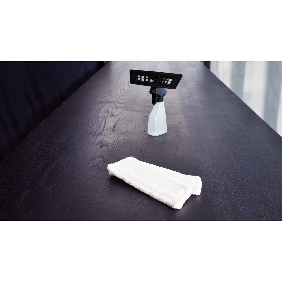 Un tavolo in legno scuro con un flacone spray per la pulizia e un tampone in microfibra bianca. Lo sfondo mostra una finestra.