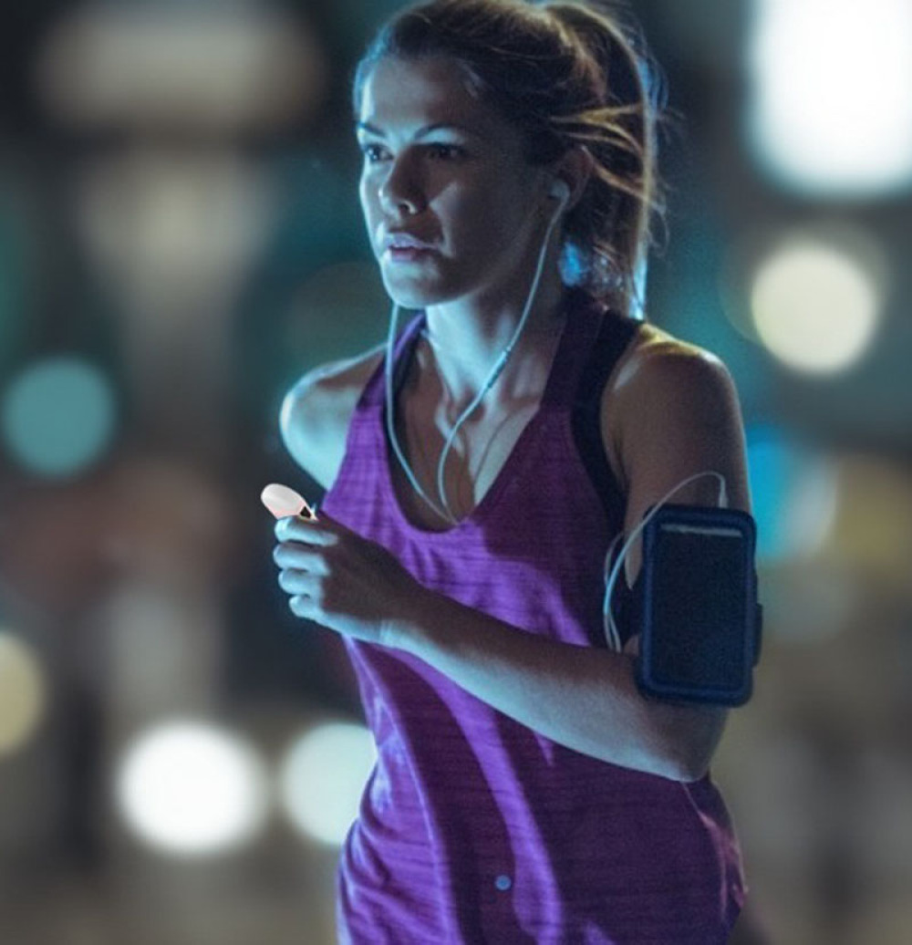 Eine Frau joggt nachts, trägt ein lila Tanktop und Kopfhörer. Sie hält ein kleines Gerät. Ein Telefon ist an ihrem Arm.
