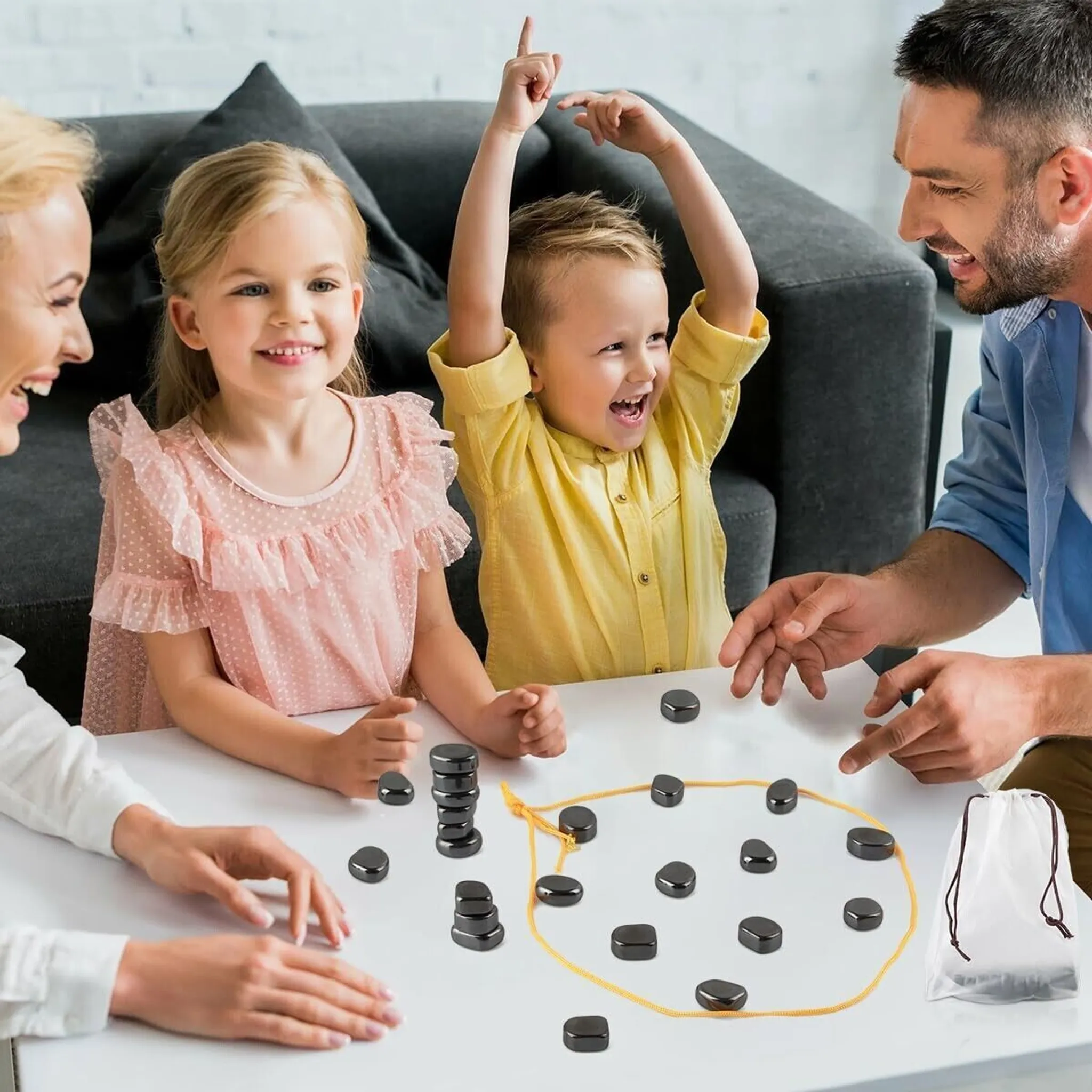 Eine Familie spielt ein Spiel mit schwarzen Steinen. Ein Junge hebt aufgeregt die Arme. Andere lächeln. Eine weiße Tasche steht daneben.