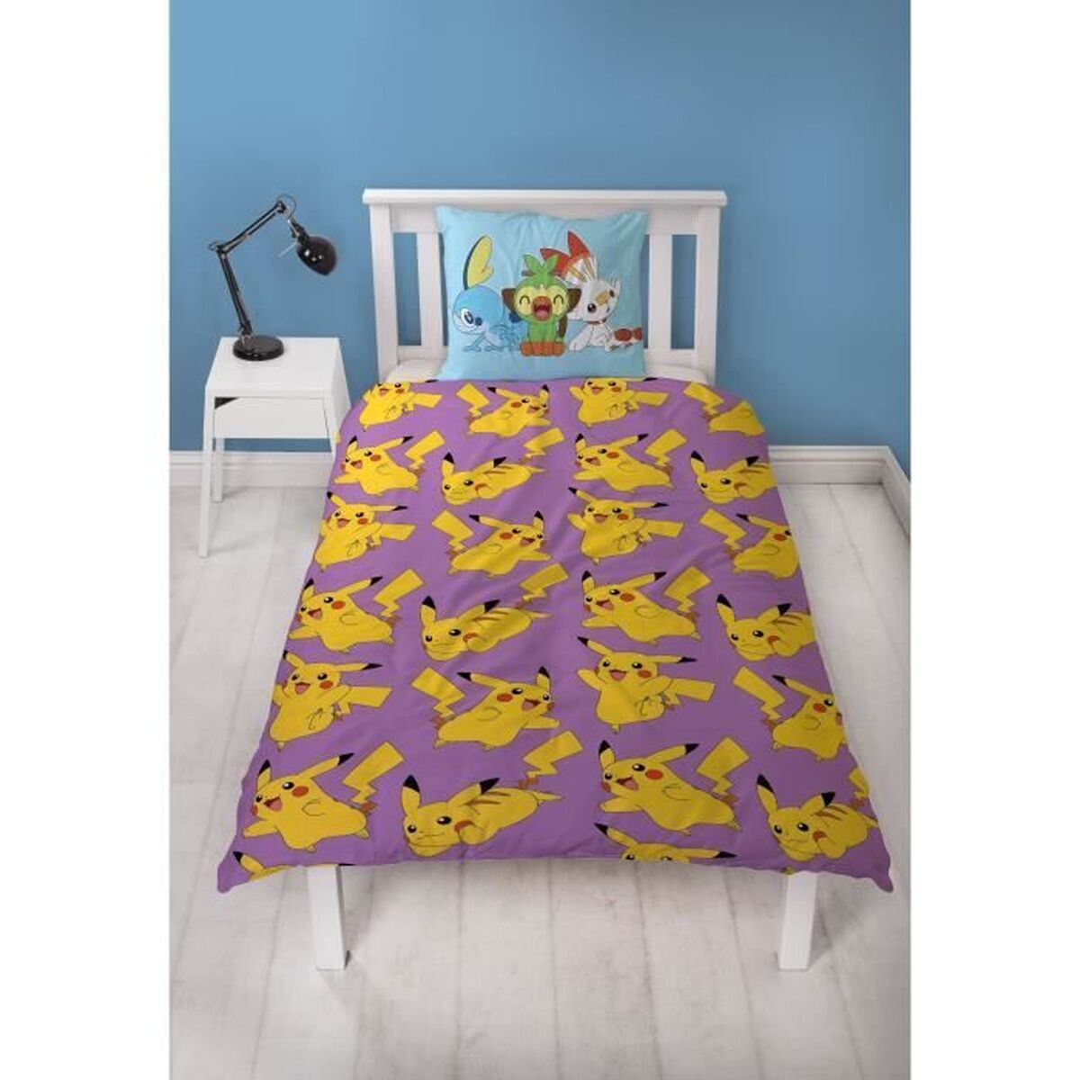 Ein Bett mit einer lila Pikachu-Bettdecke. Ein blaues Kissen hat Zeichentrickfiguren. Ein weißer Nachttisch mit einer Lampe steht neben dem Bett.