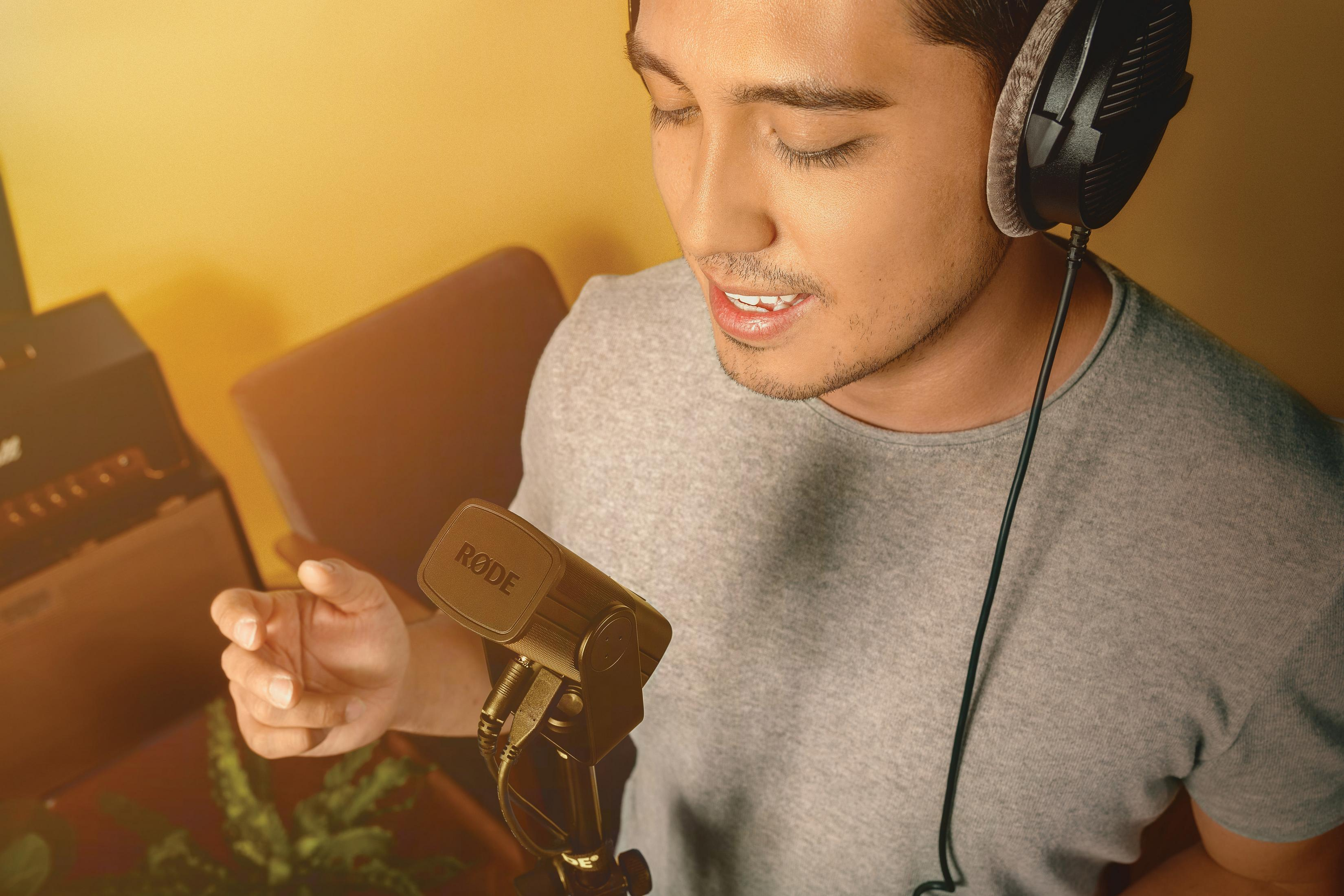 Mężczyzna w słuchawkach mówi do mikrofonu. Tło jest żółte. Ma na sobie szarą koszulę.