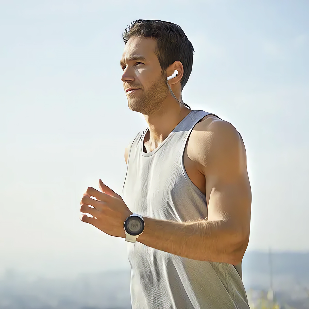 Mann rennt, trägt Ohrhörer, ein graues Tanktop und eine Smartwatch. Klarer Himmel im Hintergrund.