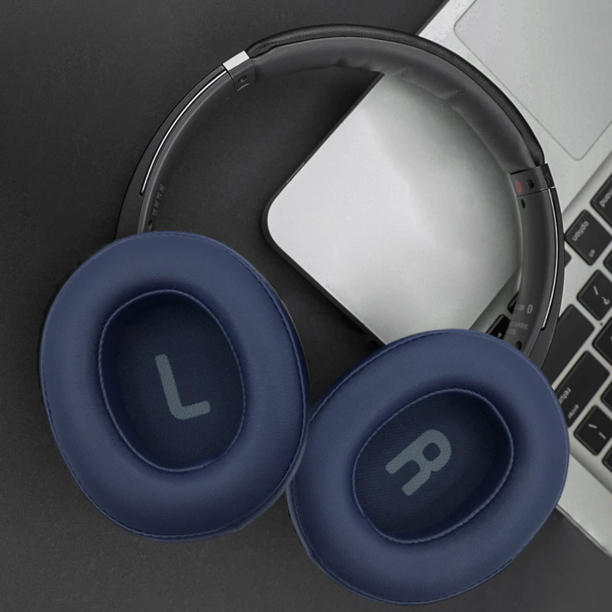 Over-Ear-Kopfhörer mit marineblauen Ohrmuscheln, beschriftet mit 'L' und 'R', liegen auf einer schwarzen Oberfläche neben einem Laptop.