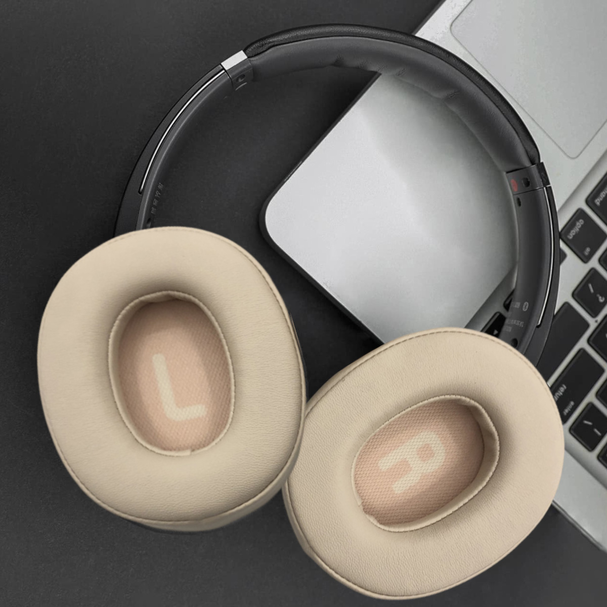 Over-Ear-Kopfhörer mit beigefarbenen Ohrmuscheln und schwarzem Bügel, teilweise auf einem Laptop und einer schwarzen Oberfläche.