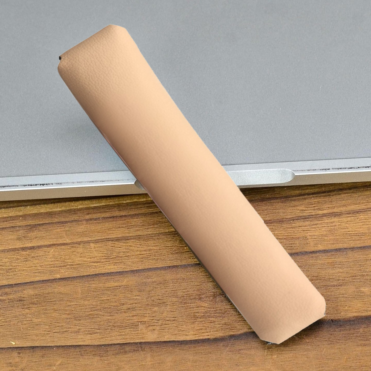 Ein beiges Lederteil liegt auf einem Laptop und einem Holztisch. Das Teil ist lang und rechteckig. Der Laptop ist silberfarben, der Tisch braun.