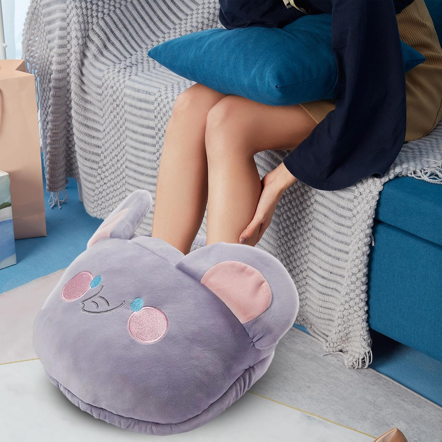 Eine Person trägt einen grauen Fußwärmer in Form eines Koalas. Die Person sitzt auf einem blauen Sofa und hält ein blaues Kissen.
