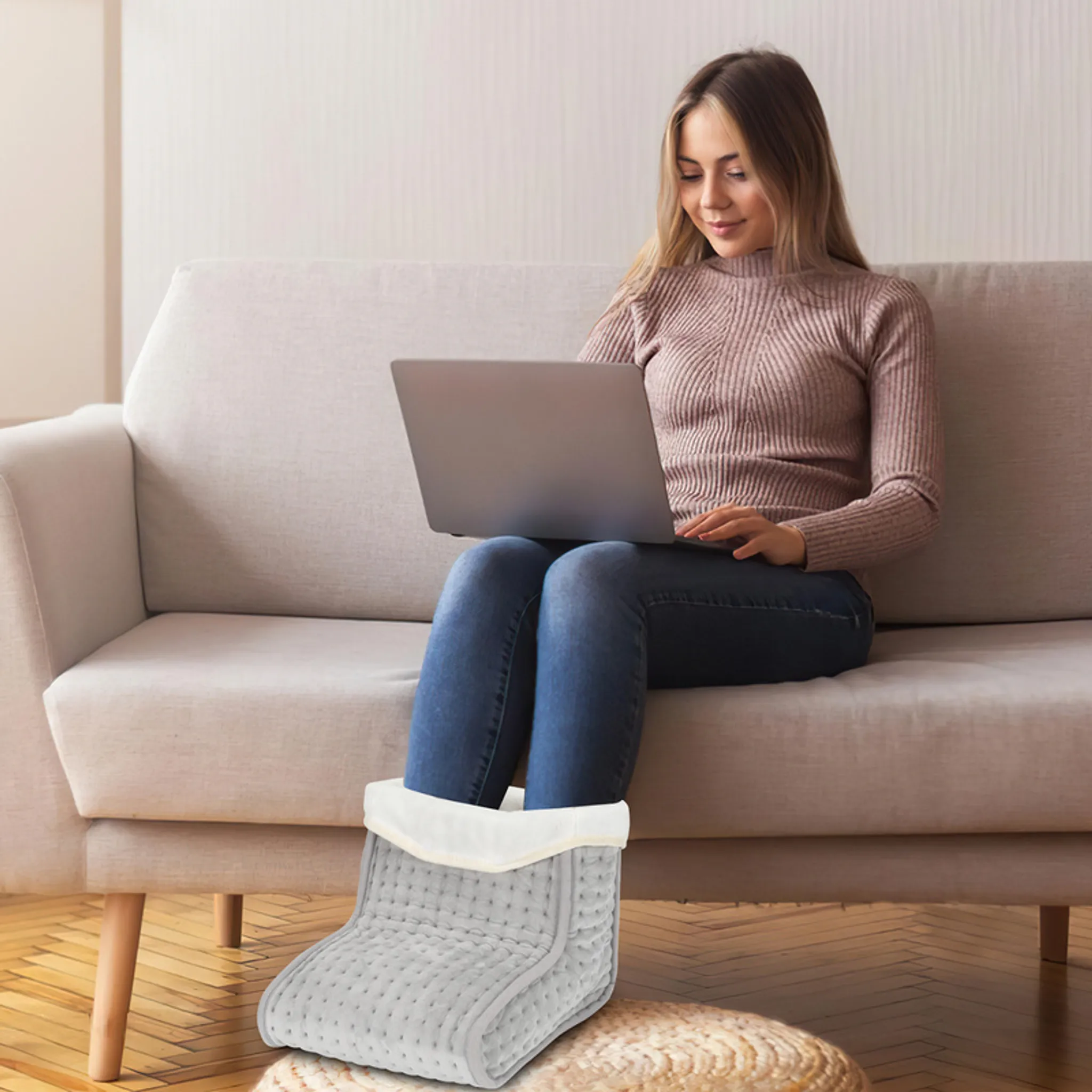 Frau auf einem Sofa, die einen Laptop benutzt, Füße in einem Fußwärmer. Beige Sofa, Jeans und ein warmer Pullover.