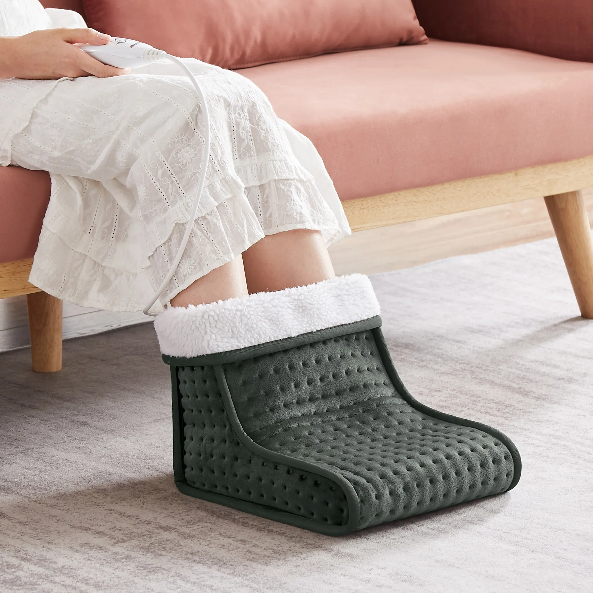 Person trägt ein weißes Kleid und einen grünen beheizten Fußwärmer. Der Hintergrund zeigt ein rosa Sofa.