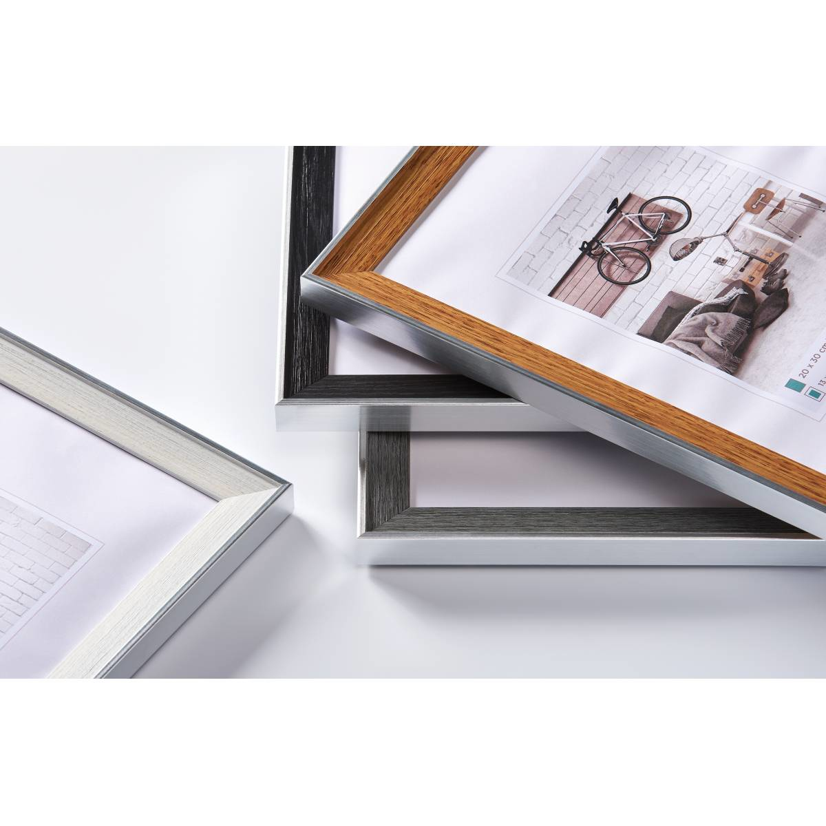 Trzy ramki na zdjęcia, srebrne i w kolorze drewna, ze zdjęciami roweru i ceglanej ściany. Białe tło.