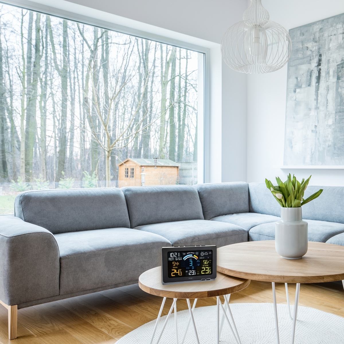 Nowoczesny salon z szarą sofą, drewnianymi stolikami i dużym oknem z widokiem na las. Na stole znajduje się stacja pogodowa.