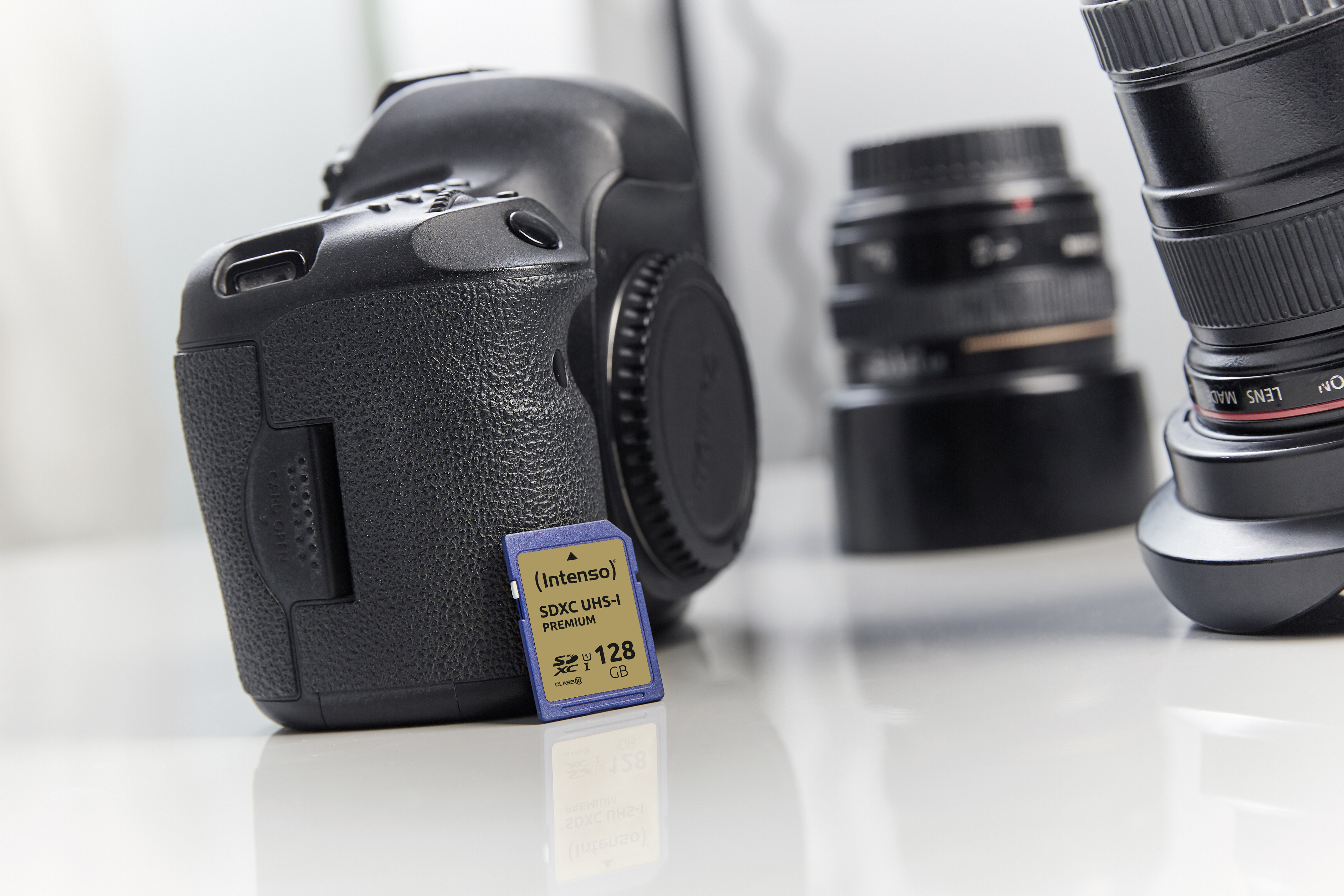 Aparat DSLR z włożoną kartą pamięci. Obiektywy rozmyte w tle. Biała, odblaskowa powierzchnia.