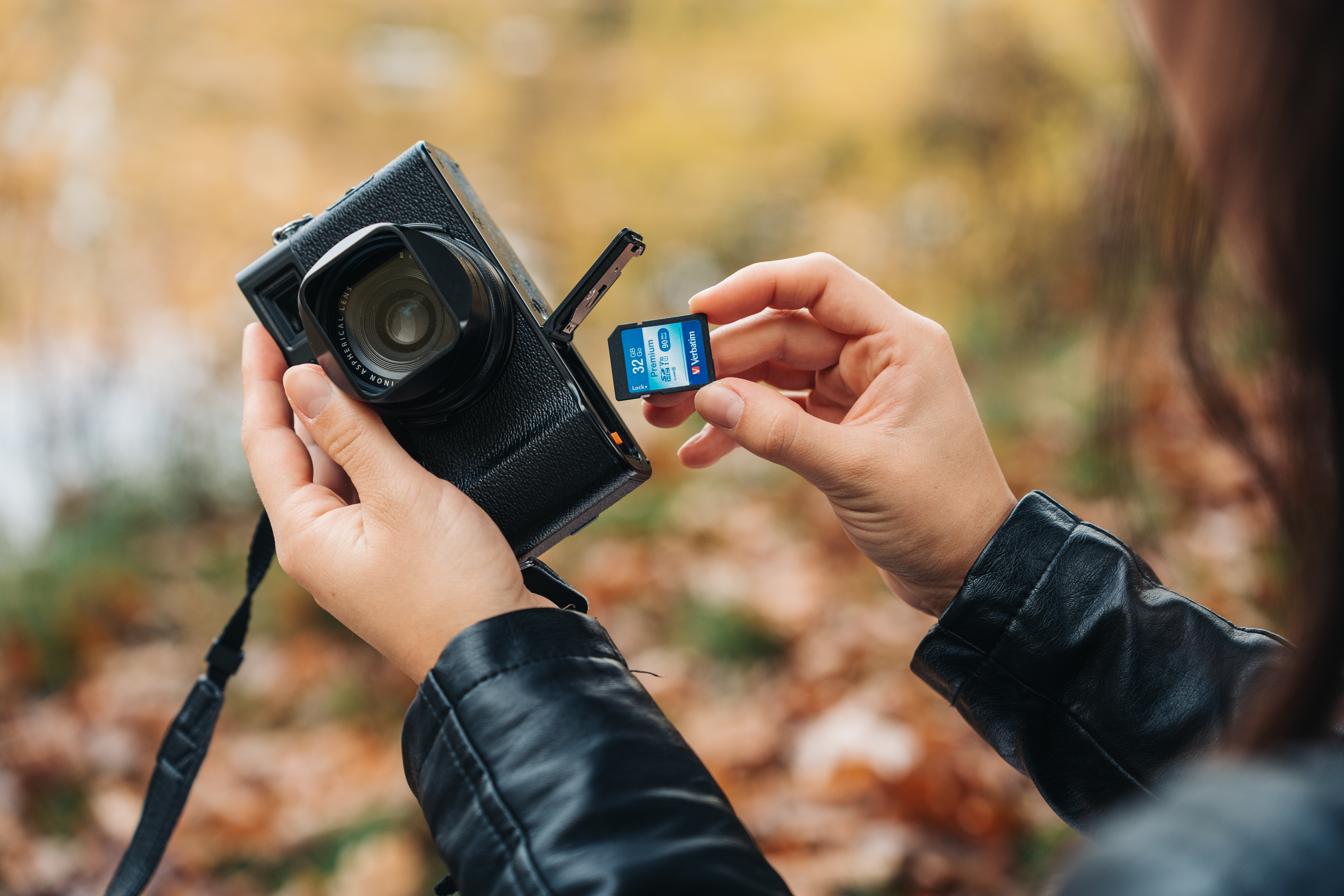 Osoba wkłada kartę SD do czarnego aparatu. Tło to rozmyte jesienne liście.