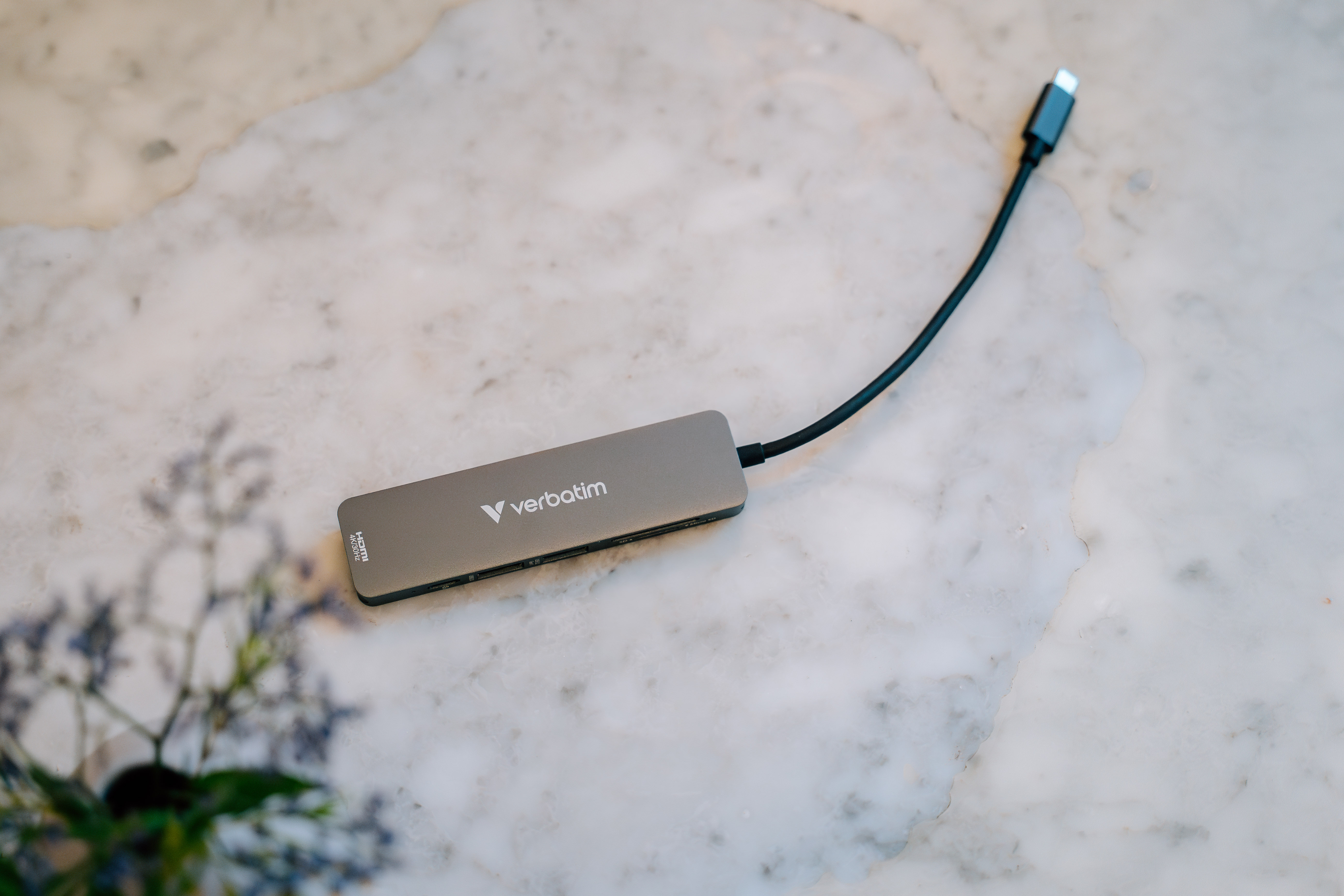 Ein grauer Verbatim USB-Hub mit schwarzem Kabel liegt auf einer Marmoroberfläche. Blumen in der linken unteren Ecke.