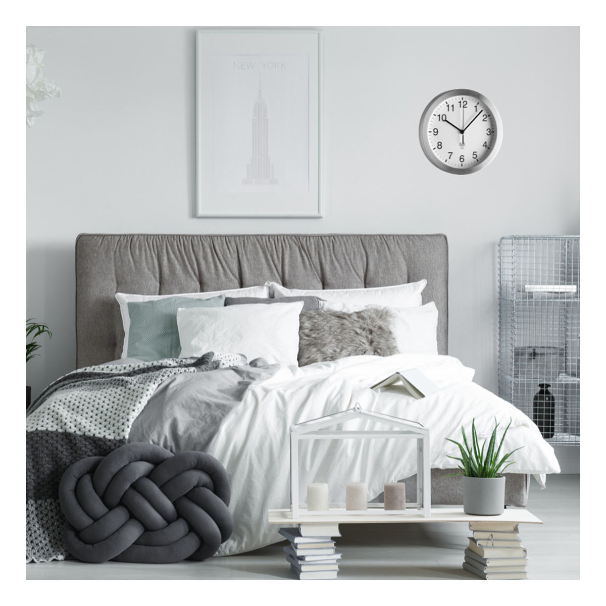 Sypialnia z łóżkiem, poduszkami, szarą ramą łóżka, zegarem i obrazem Nowego Jorku. Mały stolik ze świecami i rośliną.