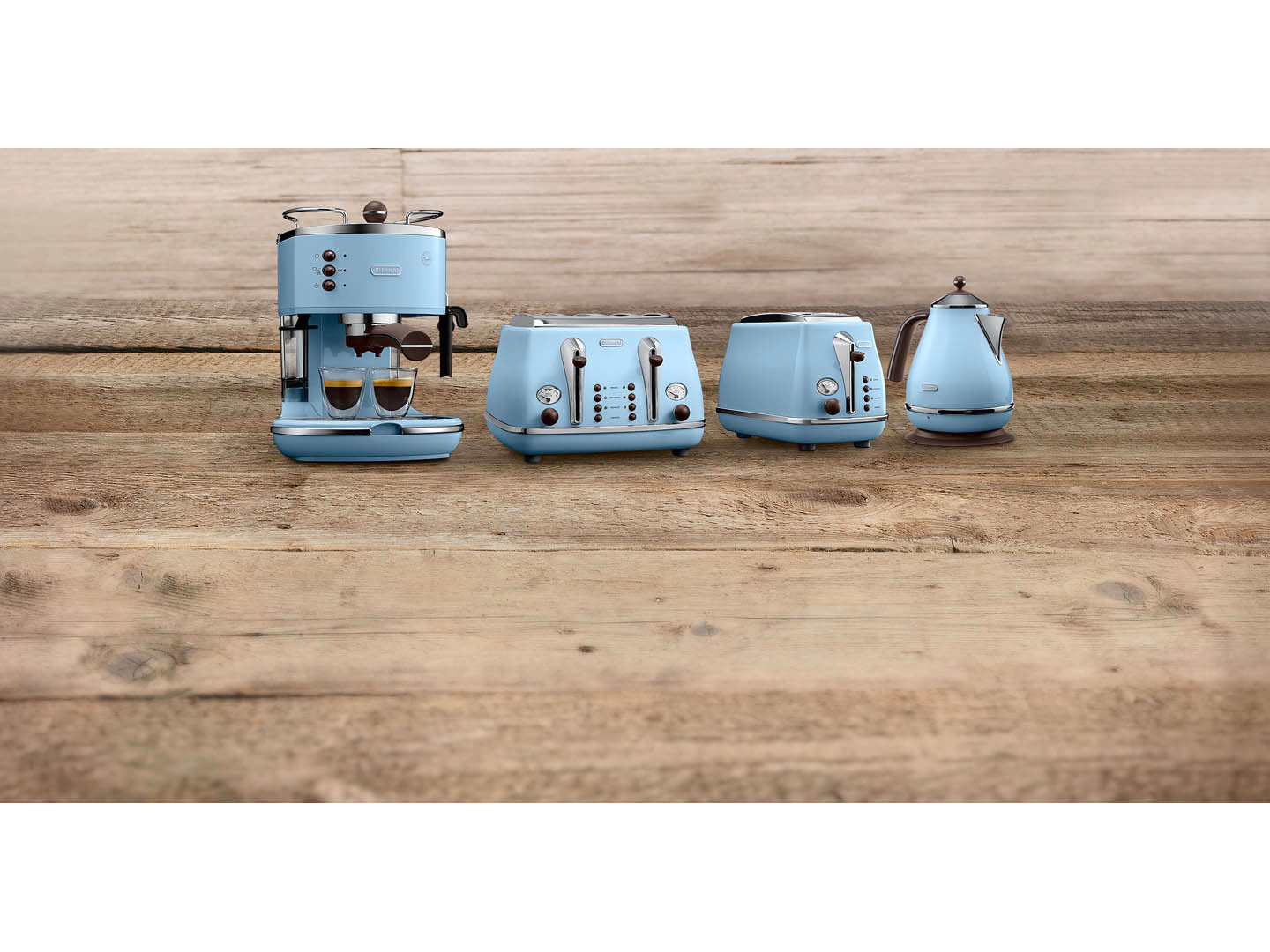 Eine hellblaue Espressomaschine, zwei Toaster und ein Wasserkocher auf einem Holztisch. Die Espressomaschine hat zwei Kaffeetassen.