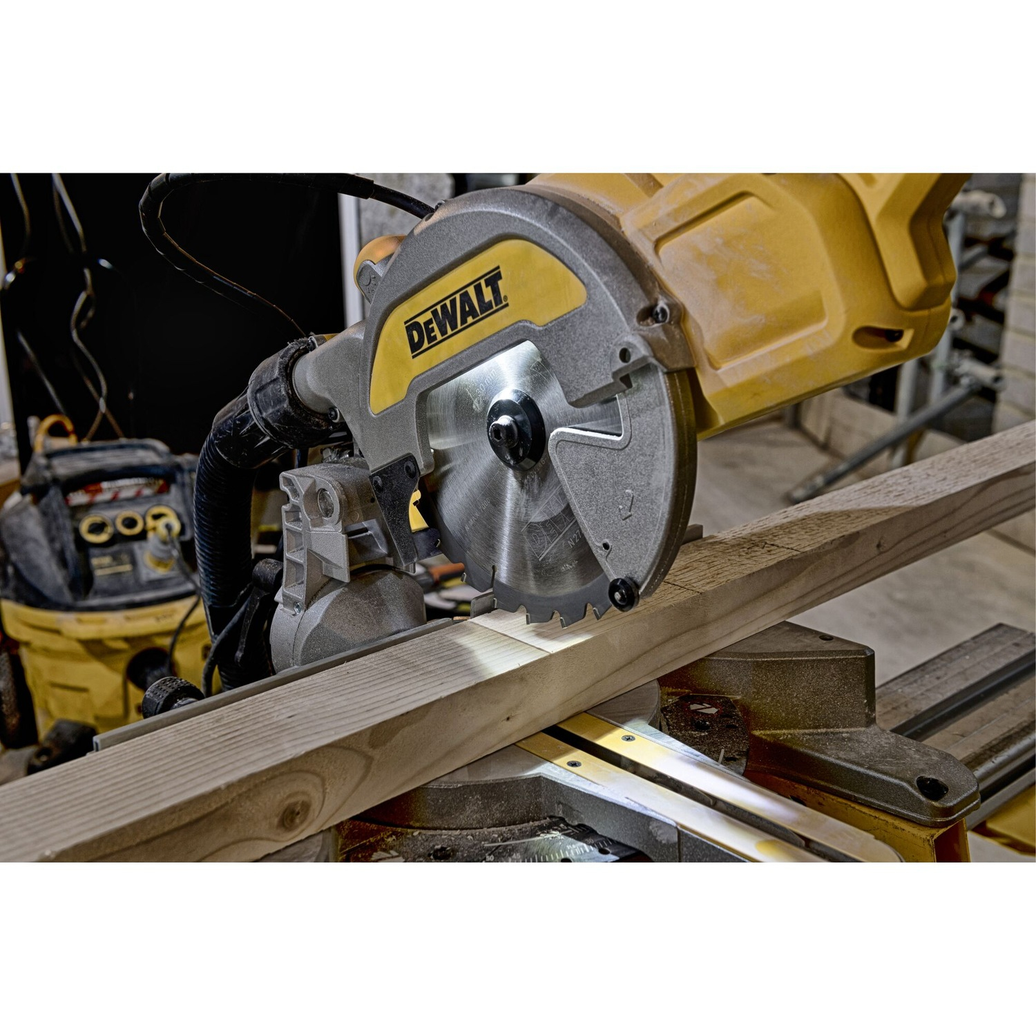 Een gele en grijze DeWalt verstekzaag snijdt een houten plank. Op de achtergrond staat een werkplaatstofzuiger.