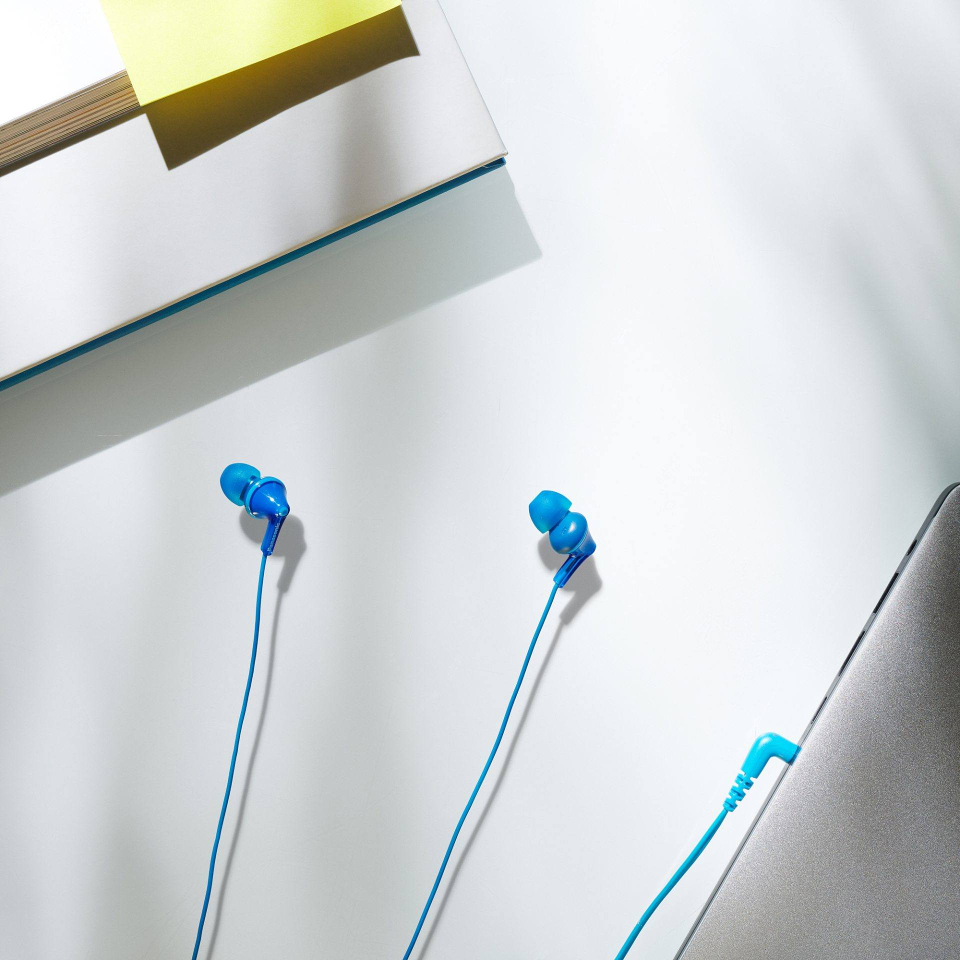 Niebieskie słuchawki z laptopem i książką z żółtą karteczką na białej powierzchni.