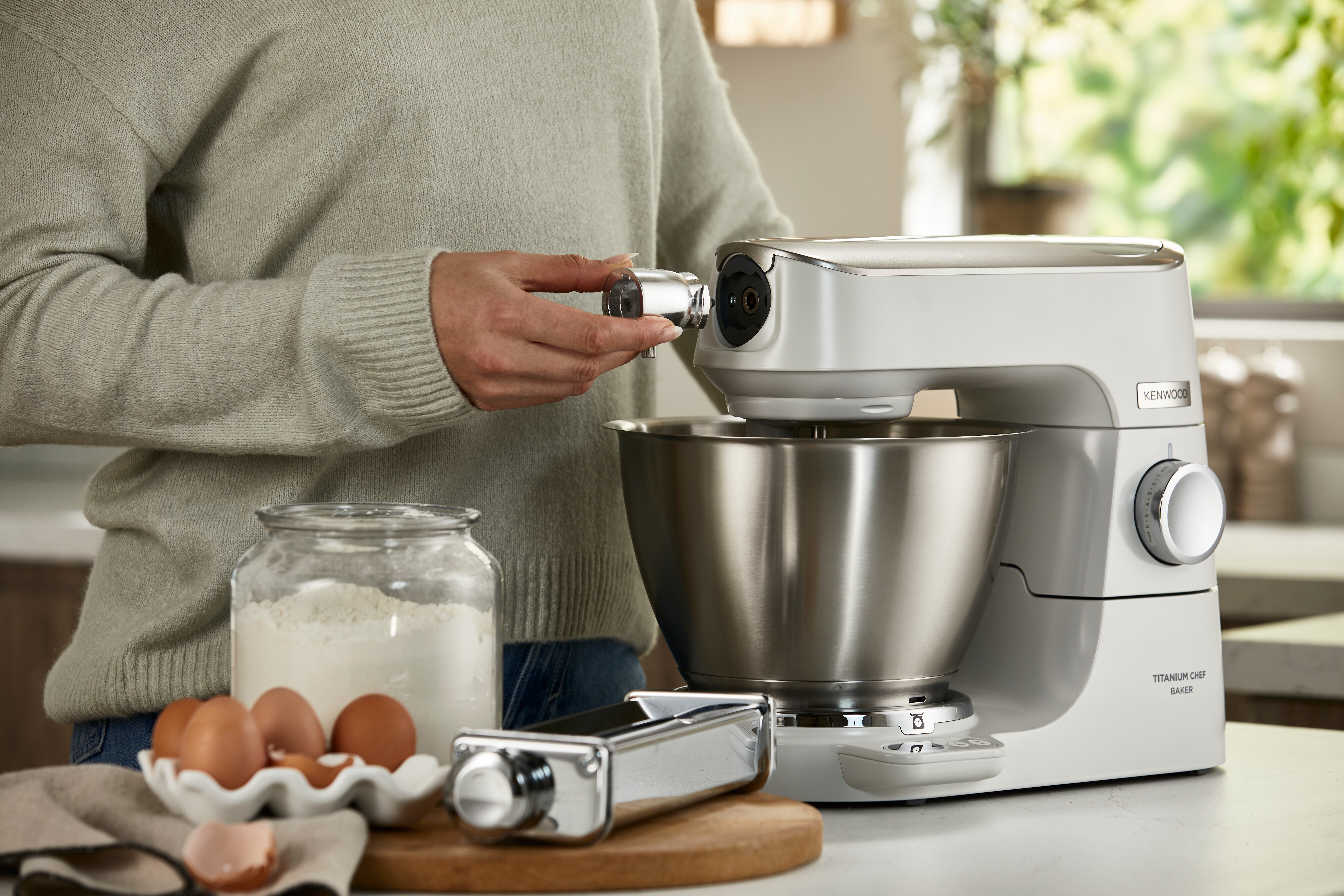 Osoba mocuje akcesorium do białego miksera. W pobliżu znajdują się składniki, takie jak jajka i mąka.