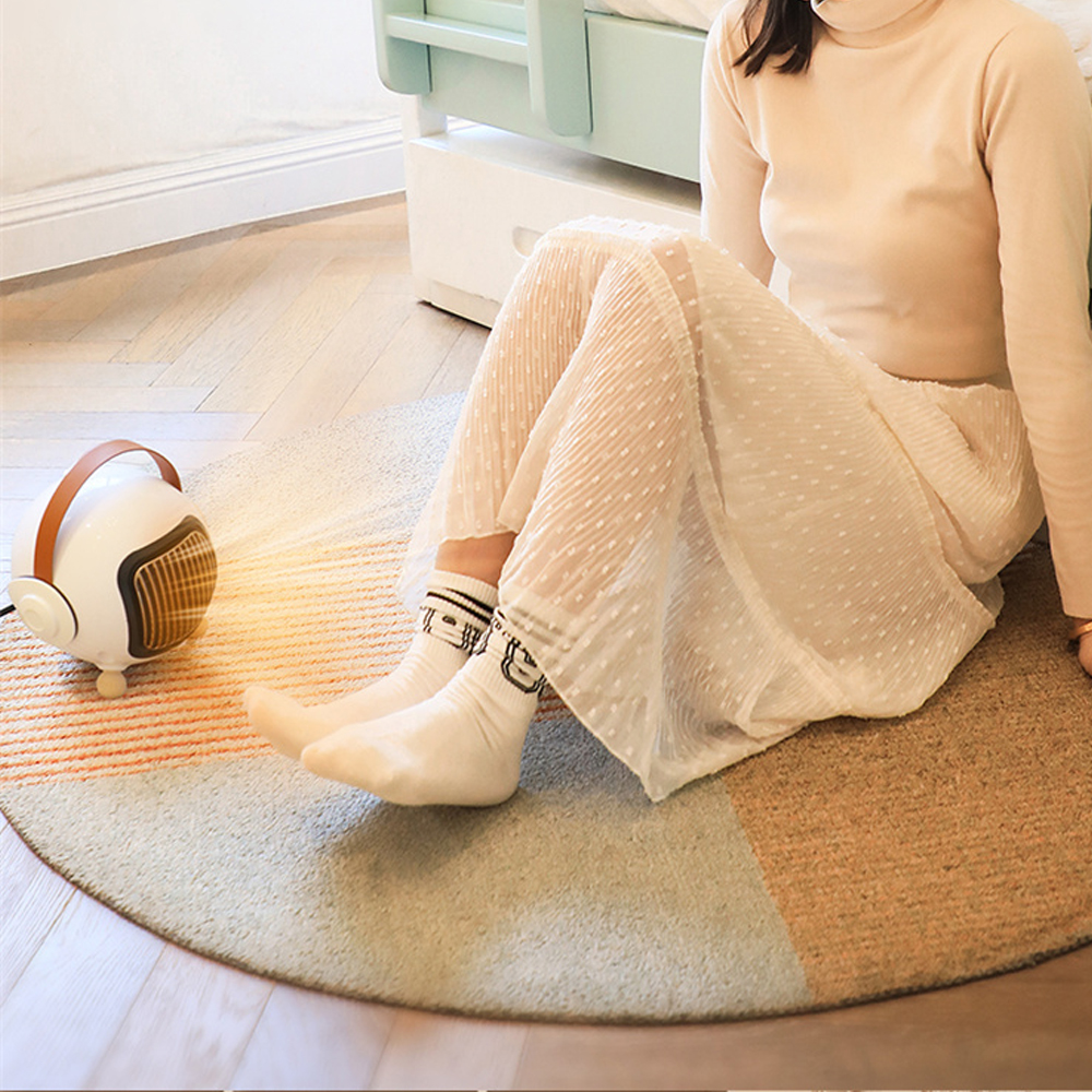 Eine Frau sitzt in der Nähe einer Heizung auf einem Teppich. Sie trägt ein cremefarbenes Oberteil, einen Rock und Socken. Die Heizung ist weiß mit braunem Griff.