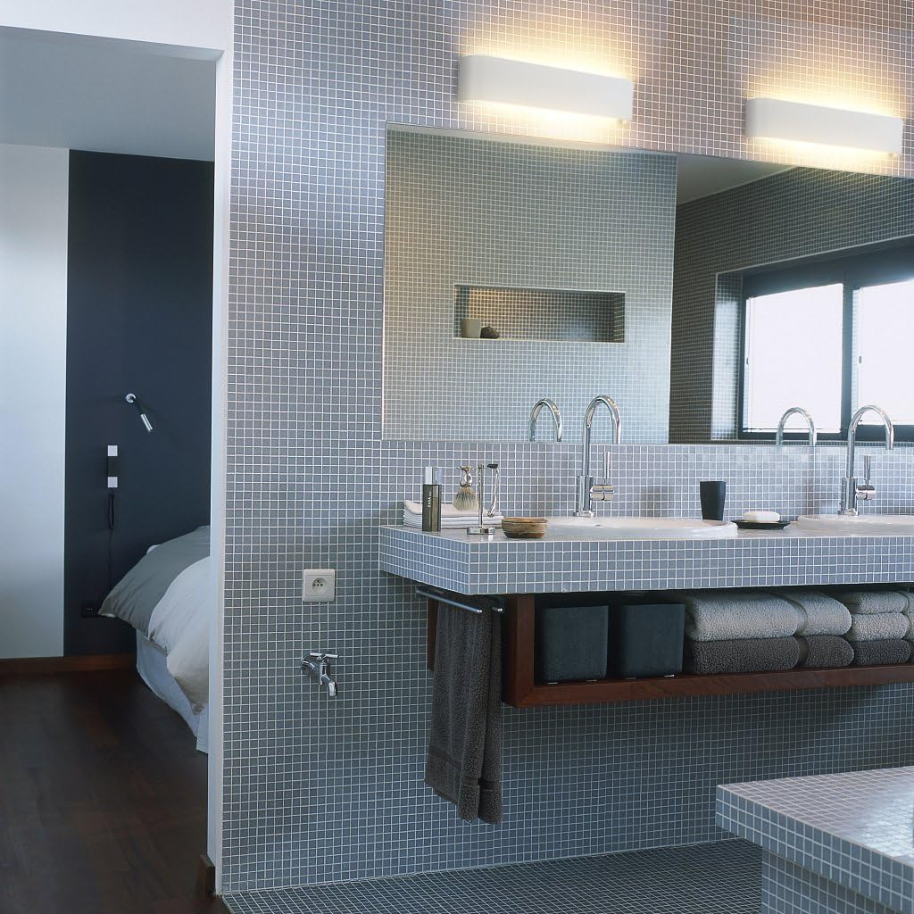 Badezimmer mit grauen Fliesen, großem Spiegel und zwei Waschbecken. Ein Handtuchhalter hält Handtücher. Ein Schlafzimmer ist durch eine Tür sichtbar.