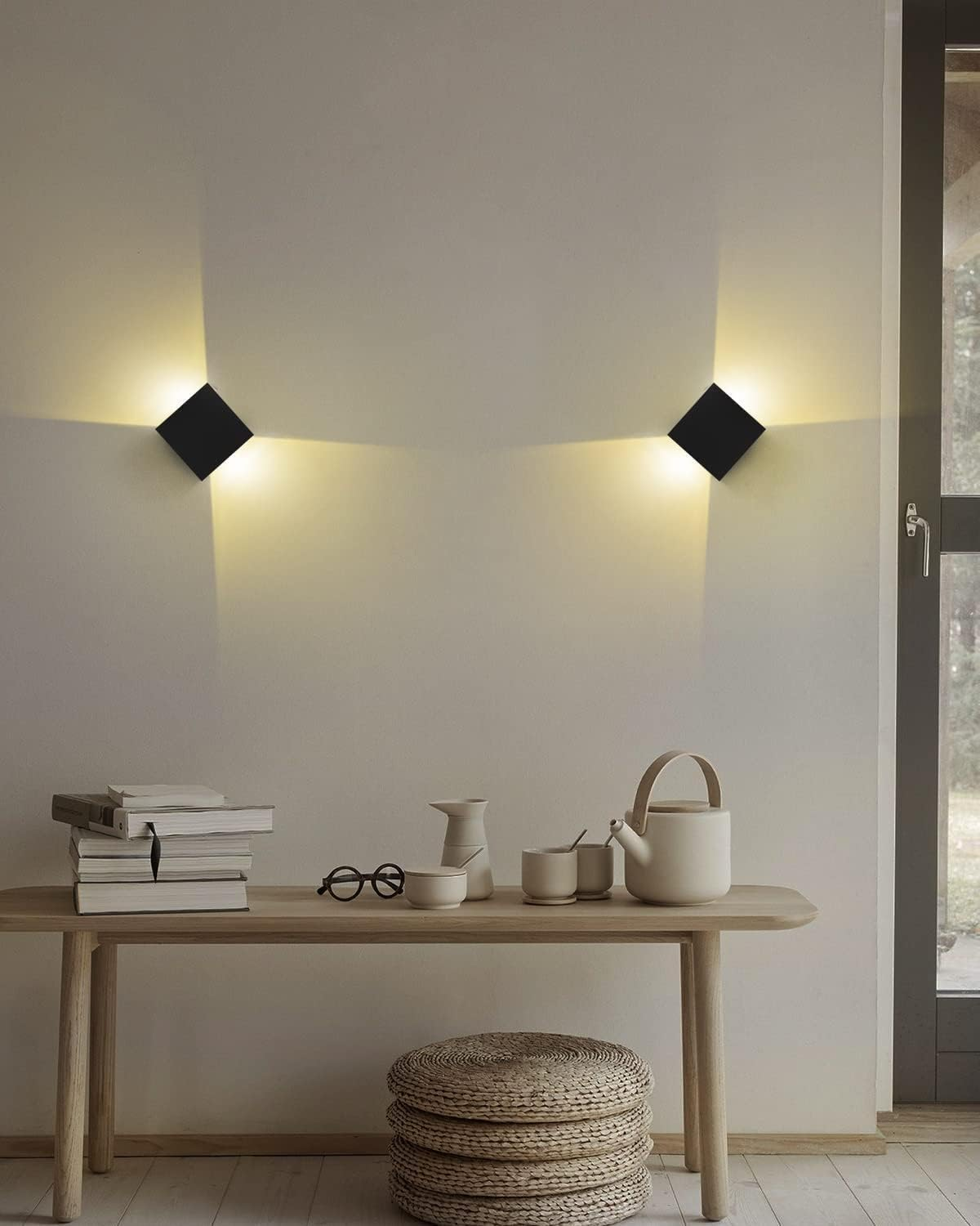 Zwei schwarze quadratische Lampen beleuchten eine Wand über einem Holztisch. Auf dem Tisch liegen Bücher, Gläser und eine Teekanne. Darunter steht ein gewebter Hocker.
