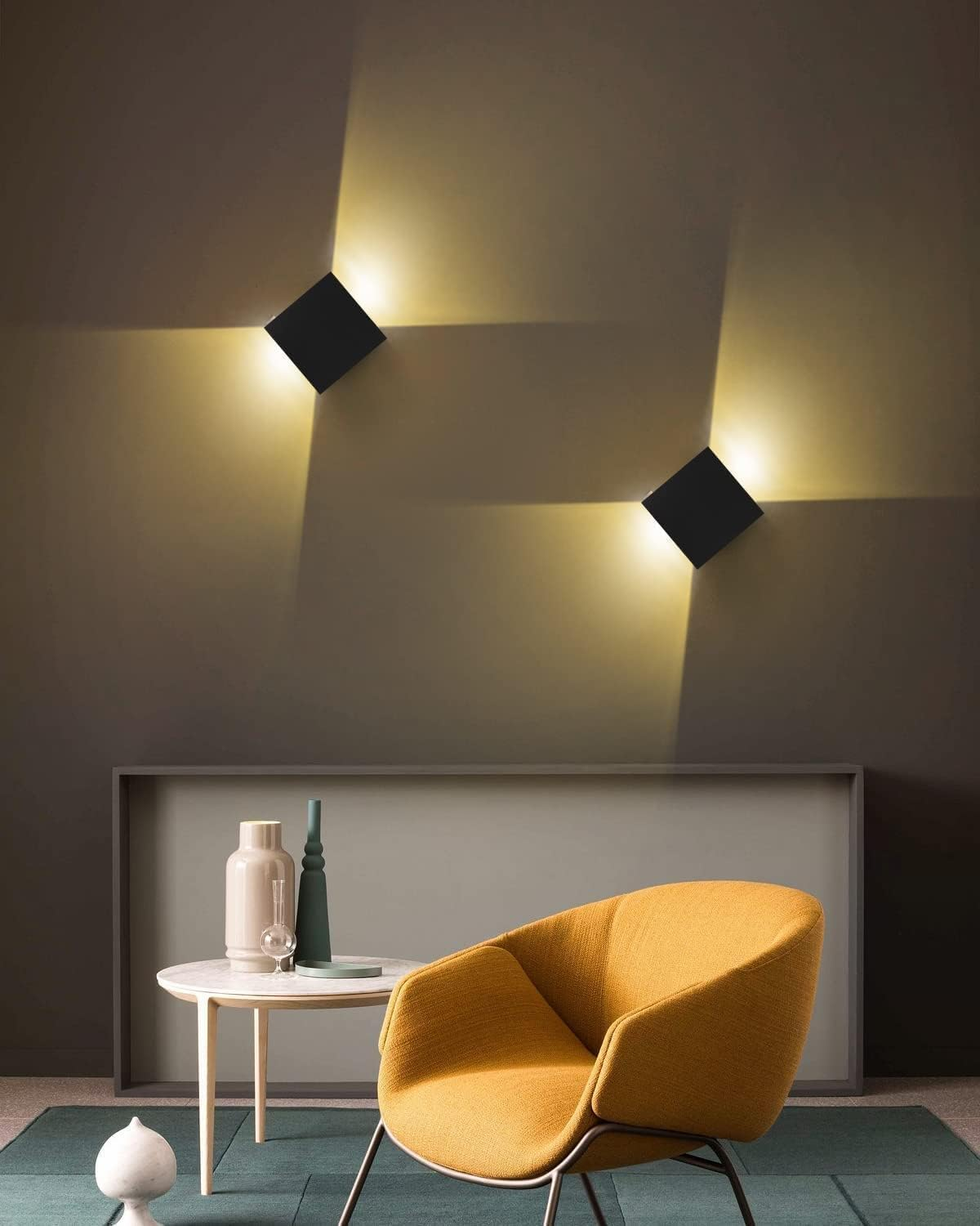 Zwei schwarze quadratische Lampen an einer grauen Wand. Ein orangefarbener Stuhl steht neben einem weißen Tisch mit Vasen. Ein grüner Teppich liegt auf dem Boden.