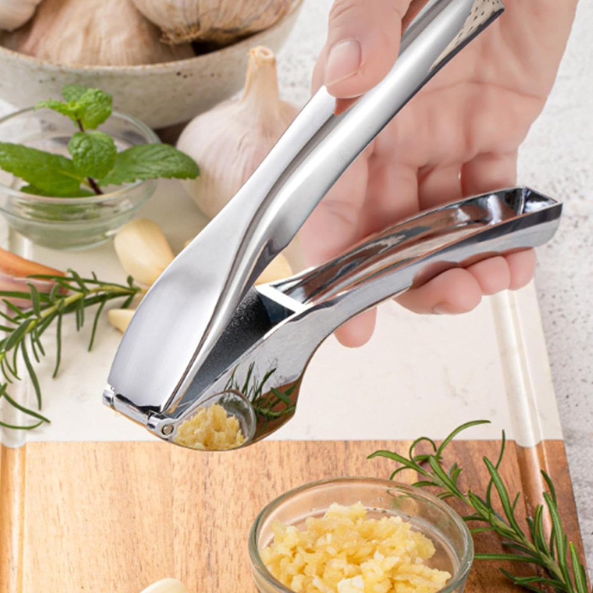 Eine Hand hält eine Knoblauchpresse über gehacktem Knoblauch in einer Schüssel. Knoblauchzehen, Kräuter und ganze Knoblauchknollen sind im Hintergrund.