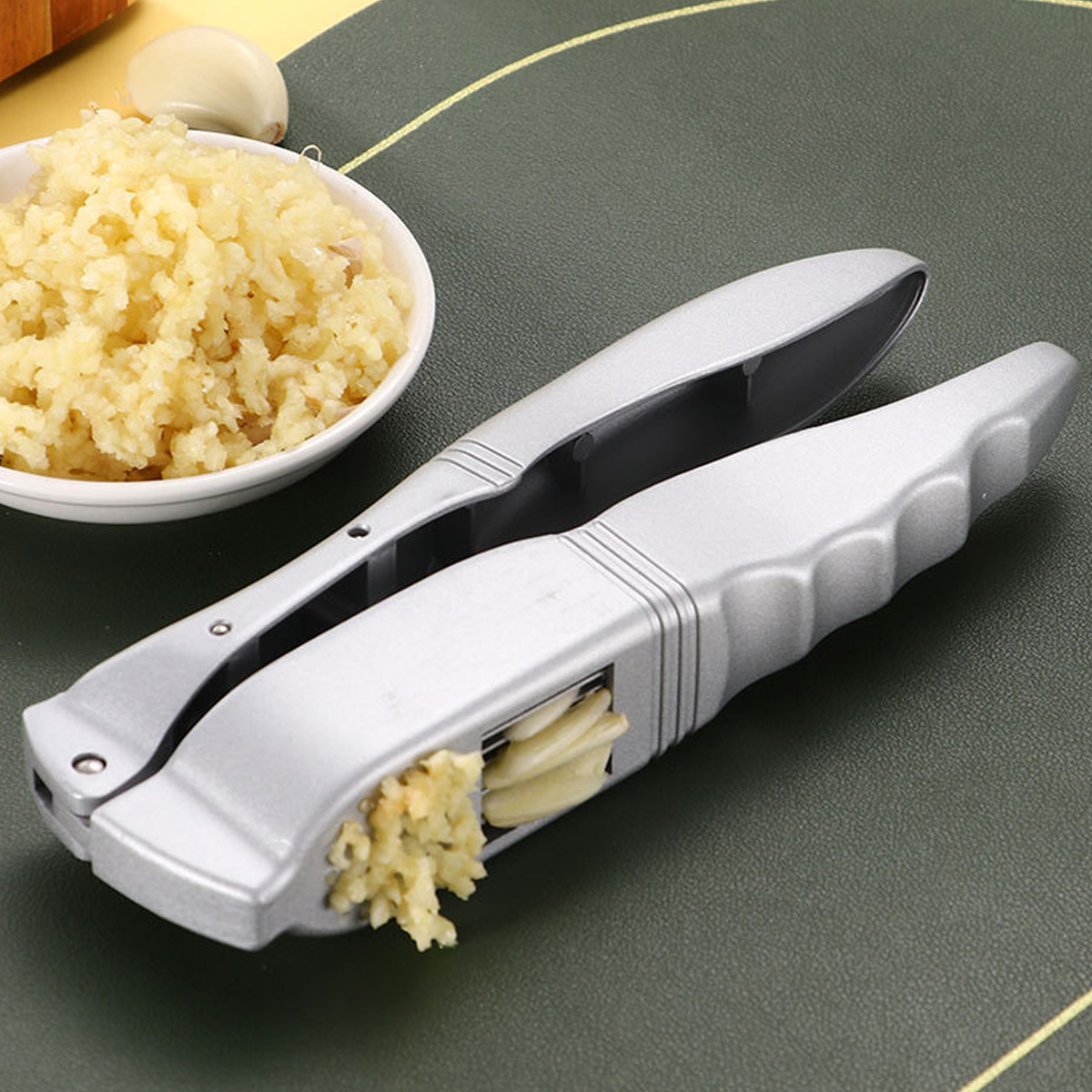 Eine silberne Knoblauchpresse mit gehacktem Knoblauch, eine Schüssel mit gehacktem Knoblauch und Knoblauchzehen auf dunklem, grünem Untergrund.