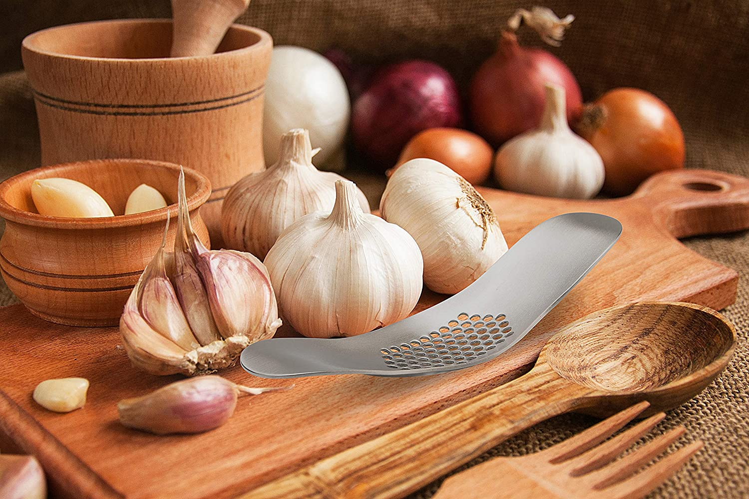 Ein Stillleben mit Knoblauch, Zwiebeln, einer Knoblauchpresse und Holzutensilien auf einem Holzbrett.