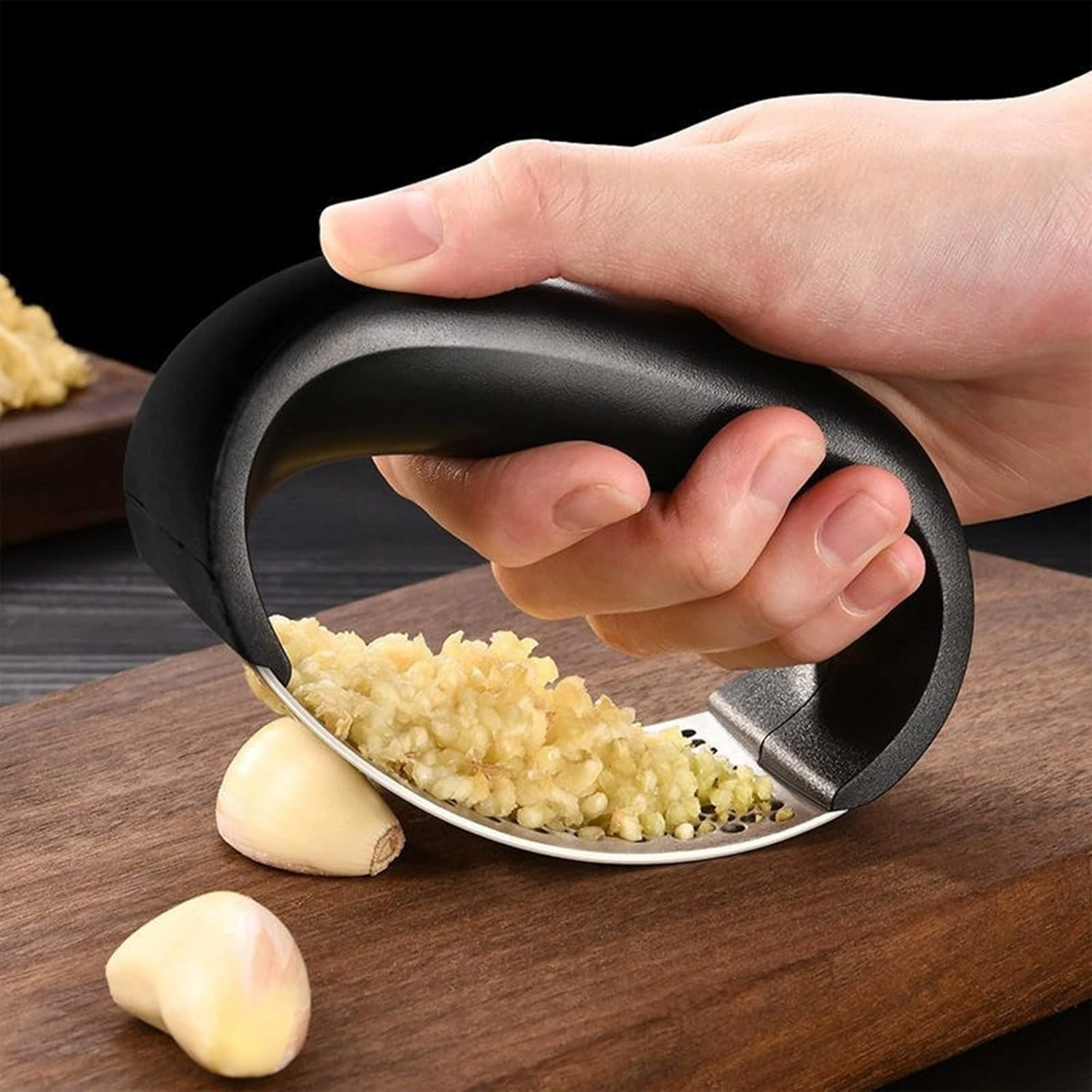 Eine Hand hält eine Knoblauchpresse und zerkleinert Knoblauch auf einem Holzbrett. Zwei Knoblauchzehen sind sichtbar. Der Hintergrund ist dunkel.