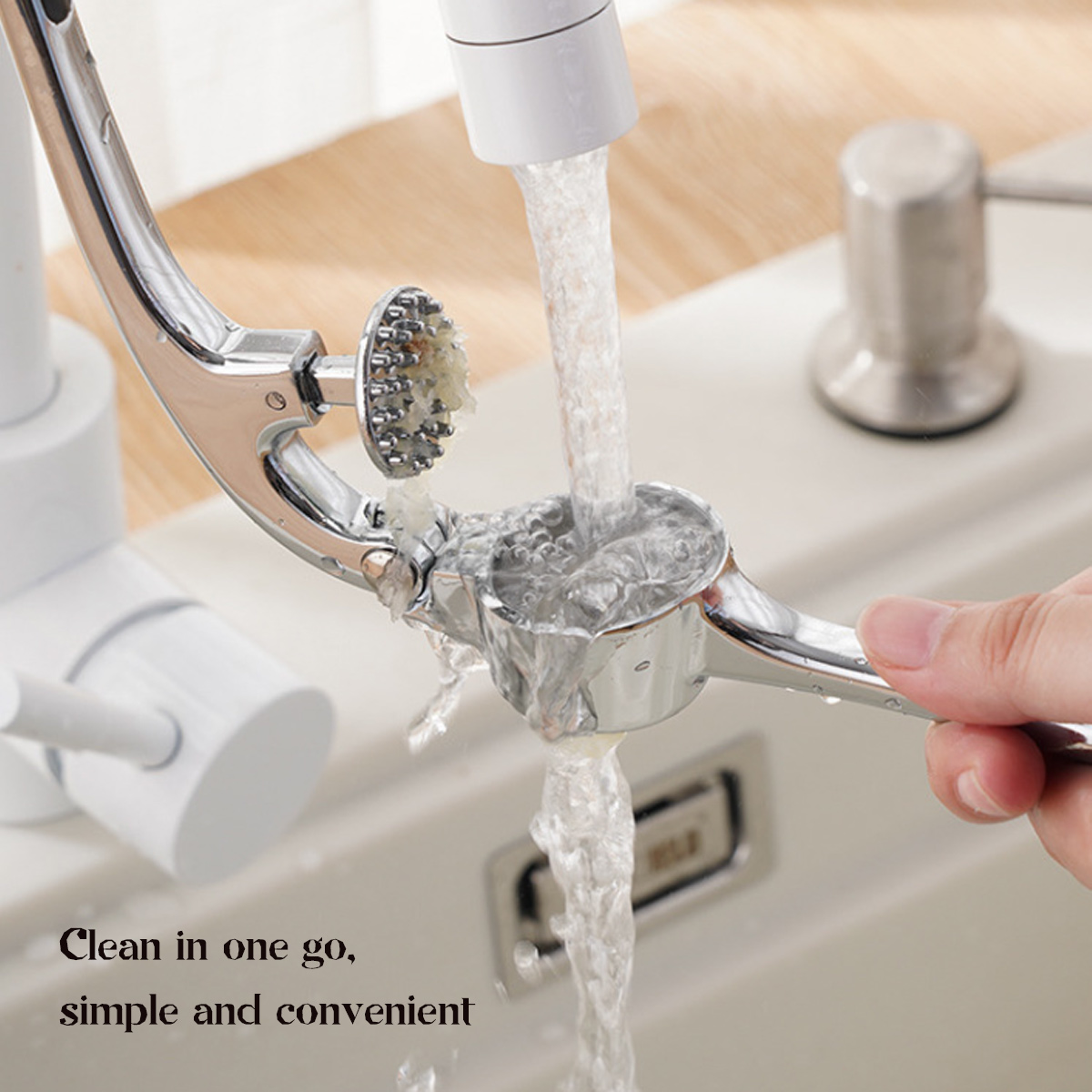Wasser fließt aus einem Wasserhahn durch eine Knoblauchpresse, die von einer Hand gehalten wird. Die Presse ist silber, der Wasserhahn ist weiß.