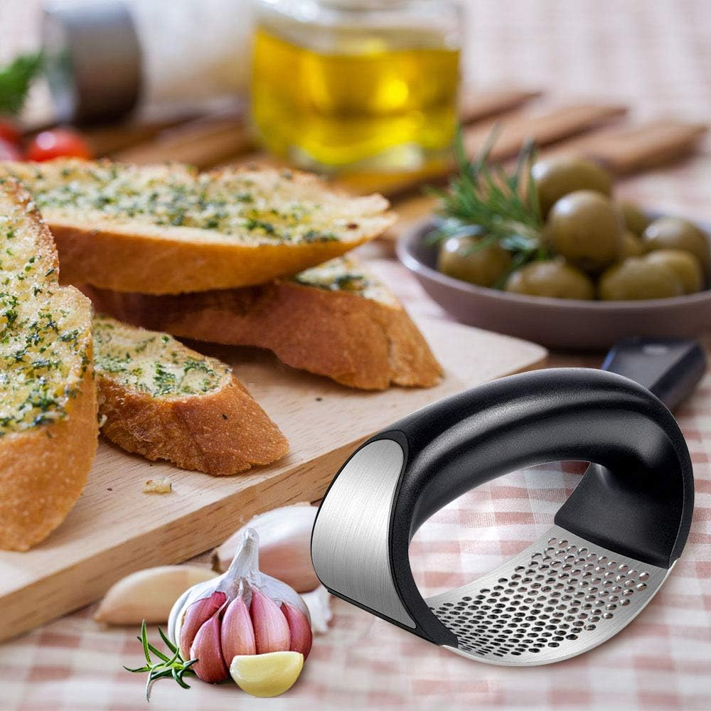 Knoblauchpresse mit Knoblauch, Brot, Öl und Oliven. Knoblauchpresse ist im Fokus, andere Artikel sind unscharf.