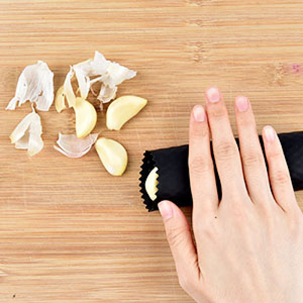 Eine Hand hält einen schwarzen Knoblauchschäler neben Knoblauchzehen und Schalen auf einem Holzschneidebrett.