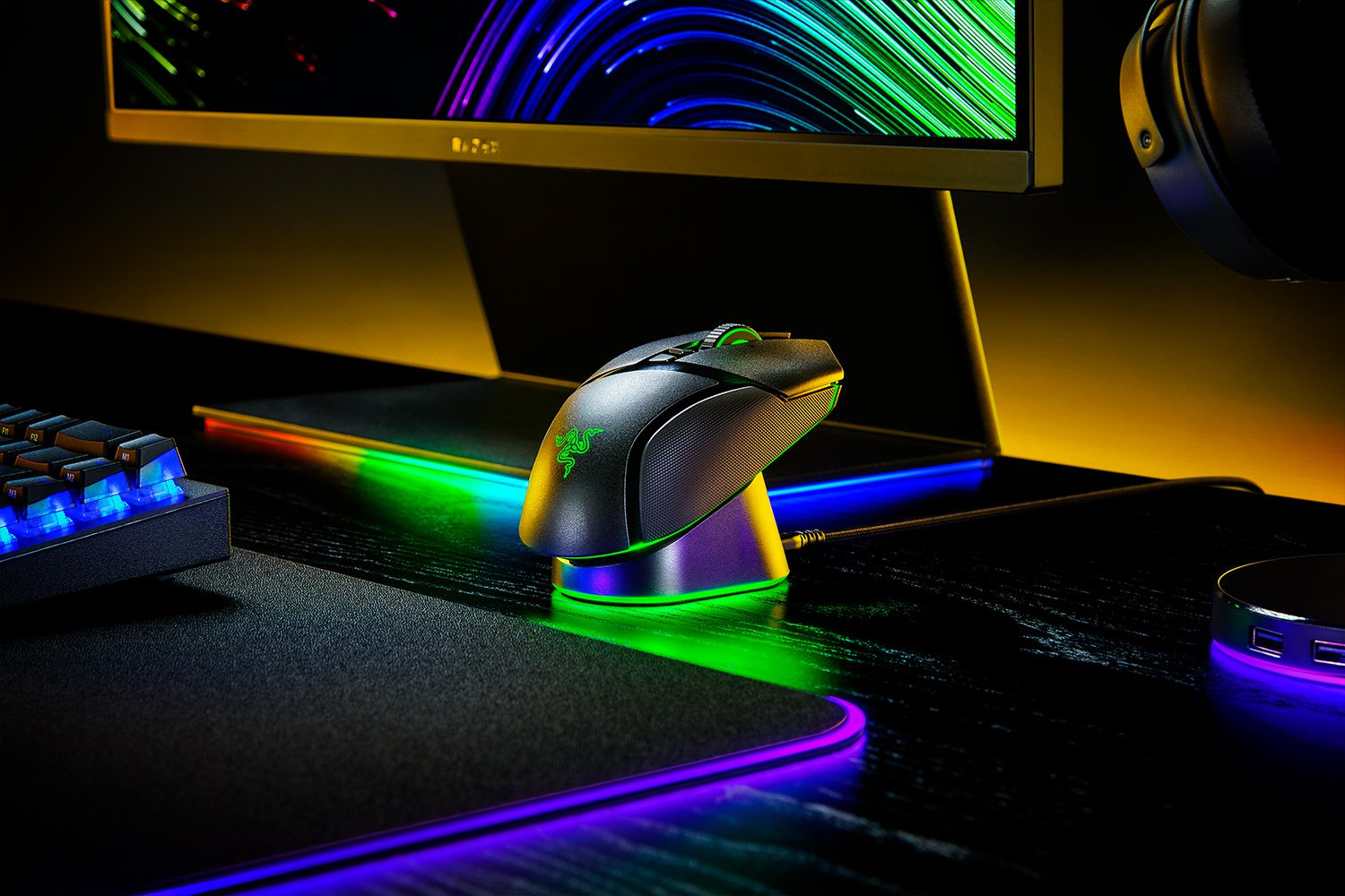 Schwarze Razer-Maus auf Ladestation, mit Tastatur und Monitor im Hintergrund. Bunte Lichter beleuchten das Setup.
