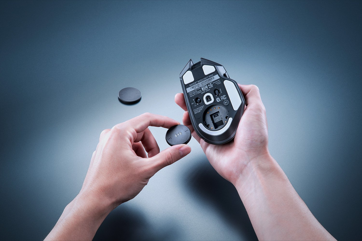 Hände halten eine schwarze Razer-Maus und einen Ladepuck. Ein zweiter Puck befindet sich im Hintergrund. Die Szene spielt sich auf grauer Oberfläche ab.