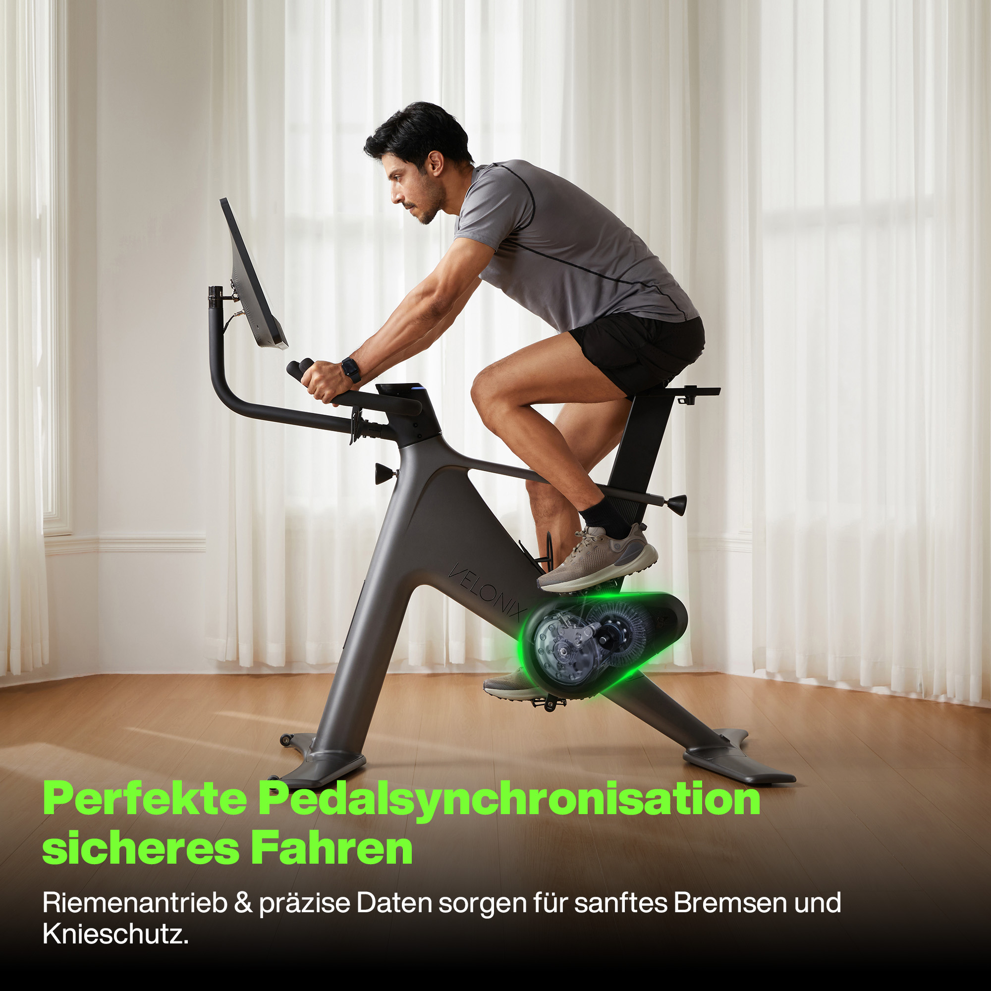 Ein Mann fährt auf einem stationären Fahrrad. Das Fahrrad ist grau. Der Mann trägt ein graues Hemd und schwarze Shorts. Der Hintergrund ist weiß.